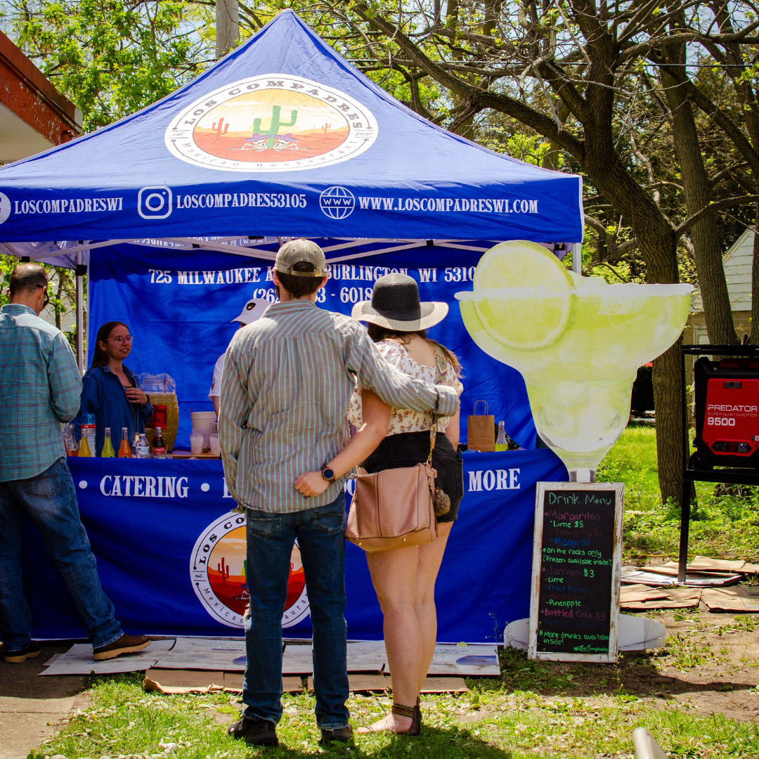 People standing at a food stand under a blue canopy with a cactus logo, ordering drinks at an outdoor event in a park with trees. Signage advertises margaritas, lime, and other beverages.