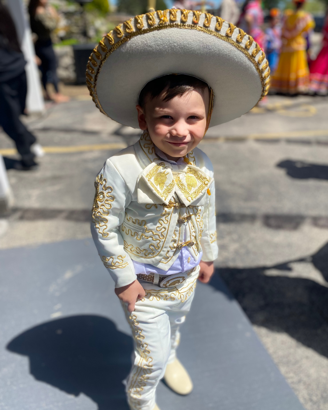 A young boy dressed in traditional Mexican charro attire, wearing a large sombrero with gold detailing and a matching white and gold embroidered suit, smiling at the camera during a cultural event.