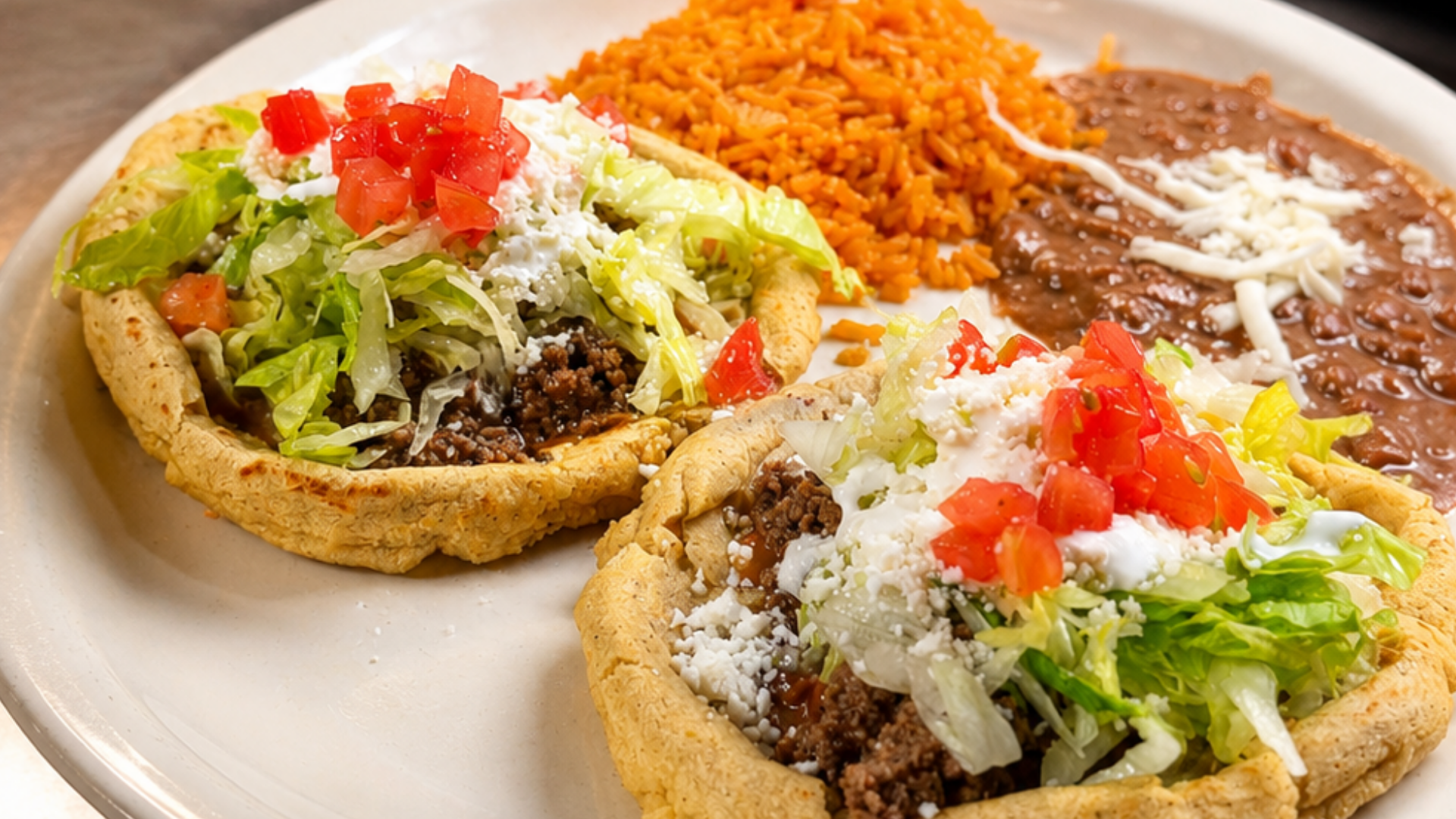 Two open taco shells filled with ground beef, shredded cheese, shredded lettuce, diced tomatoes, and sour cream, served on a white plate with Mexican rice and refried beans topped with cheese.