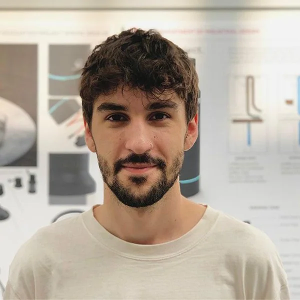 Young man with dark, wavy hair and a beard, wearing a white t-shirt, standing indoors with a blurred poster or presentation in the background.