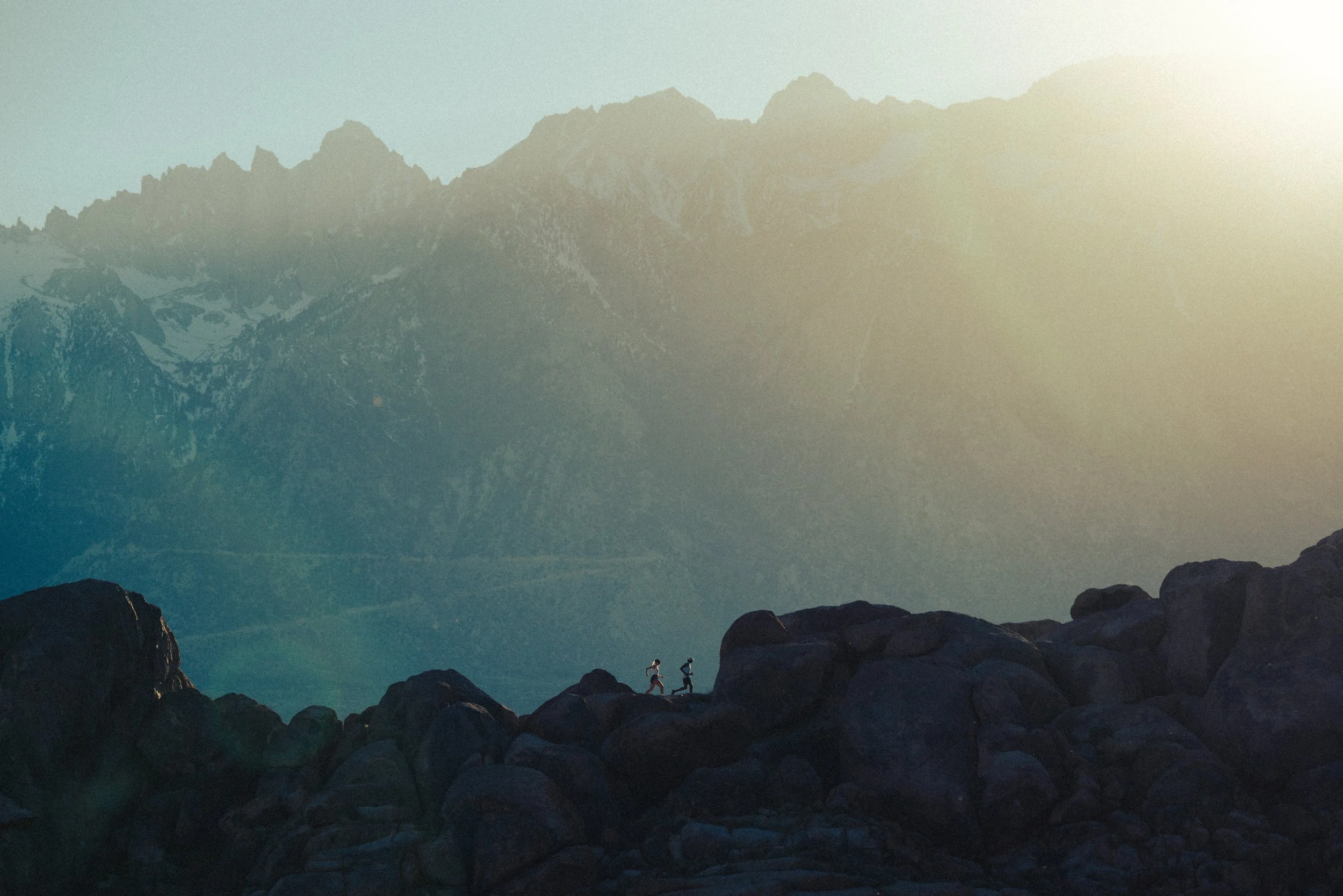 Two people hiking on rocky terrain with mountains in the background during sunset or sunrise.