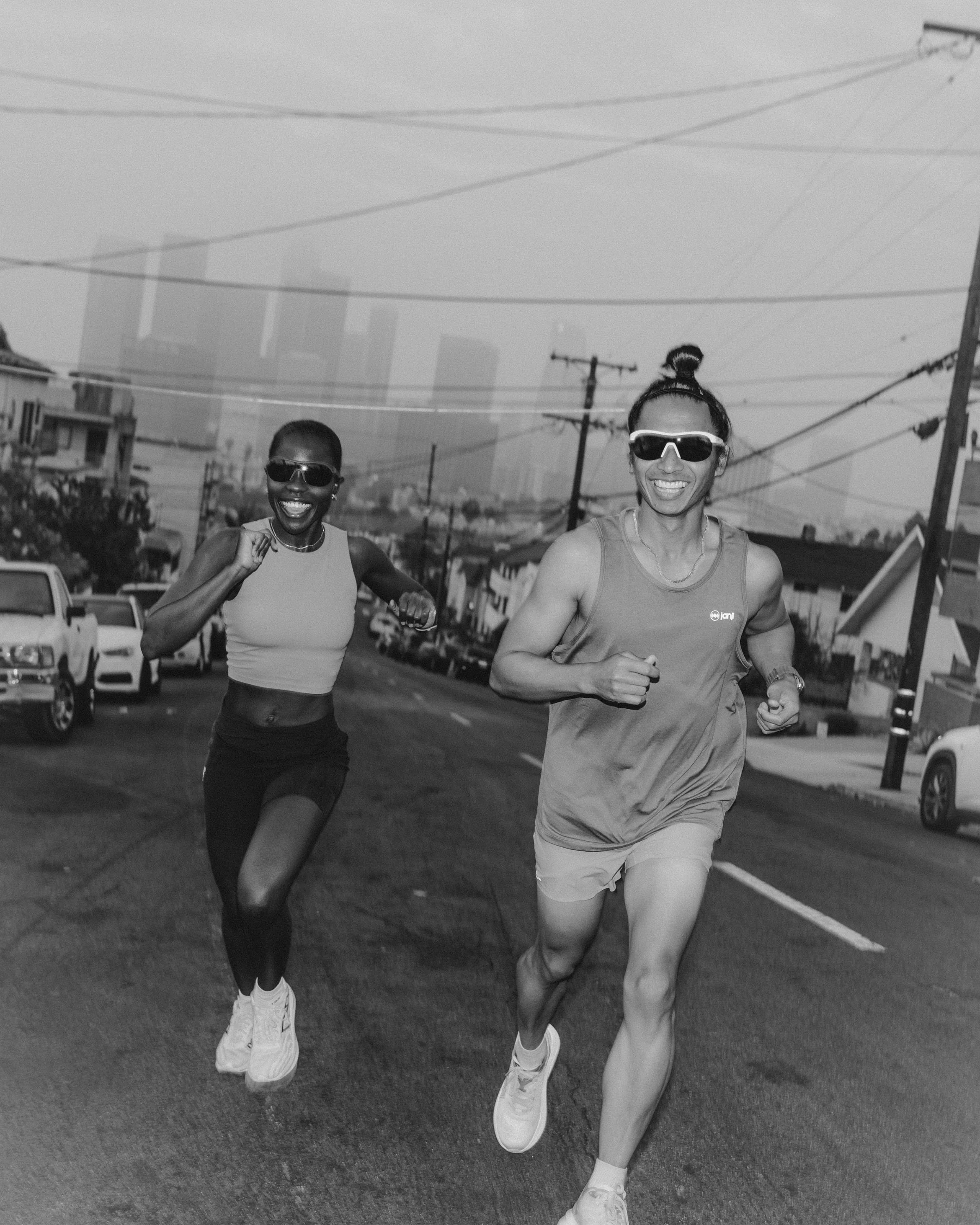 Two women running on a city street, smiling and wearing sunglasses, with cars parked along the side and power lines overhead.