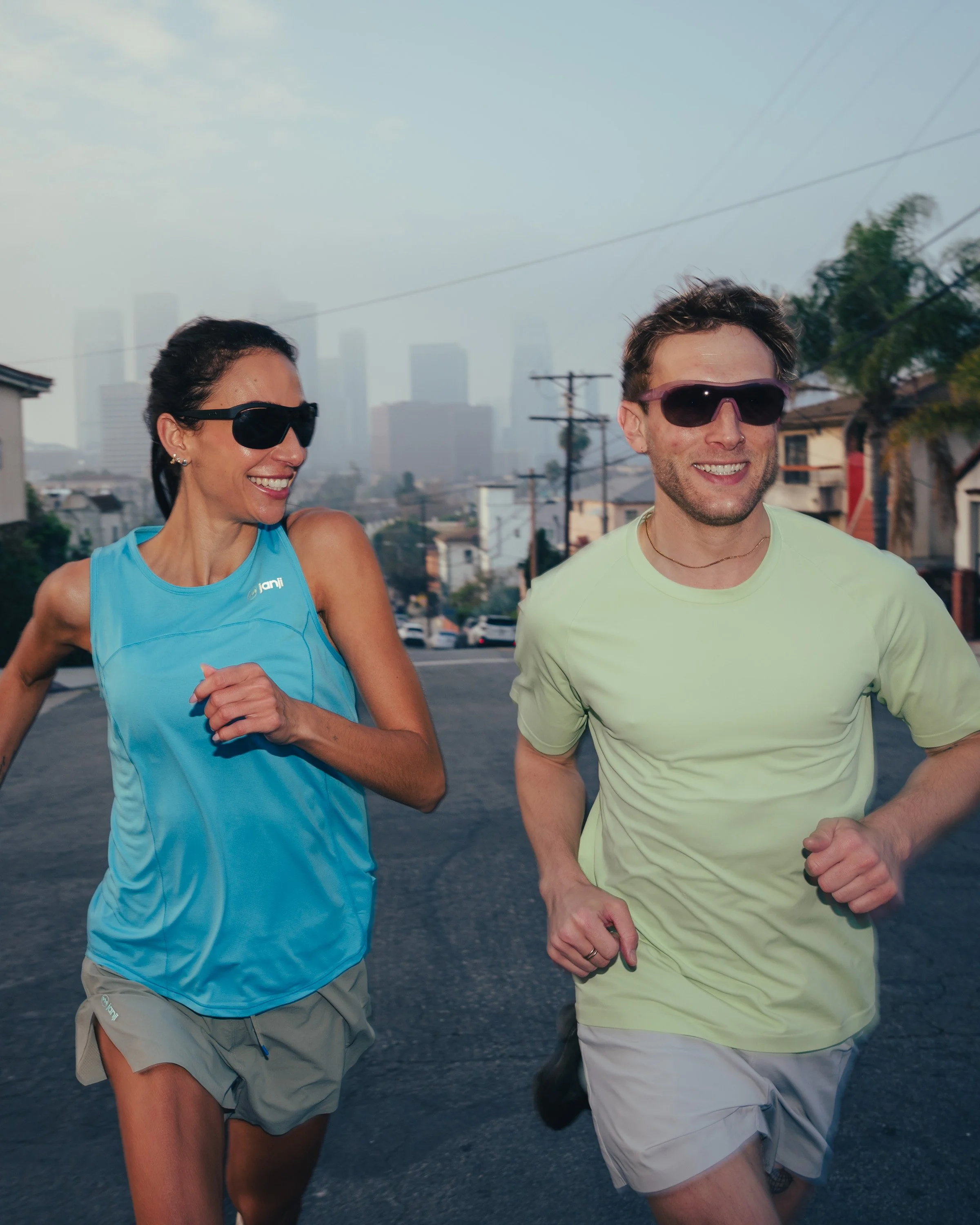 Two people running outdoors on a street with city skyline in the background, both wearing sunglasses and athletic clothing.