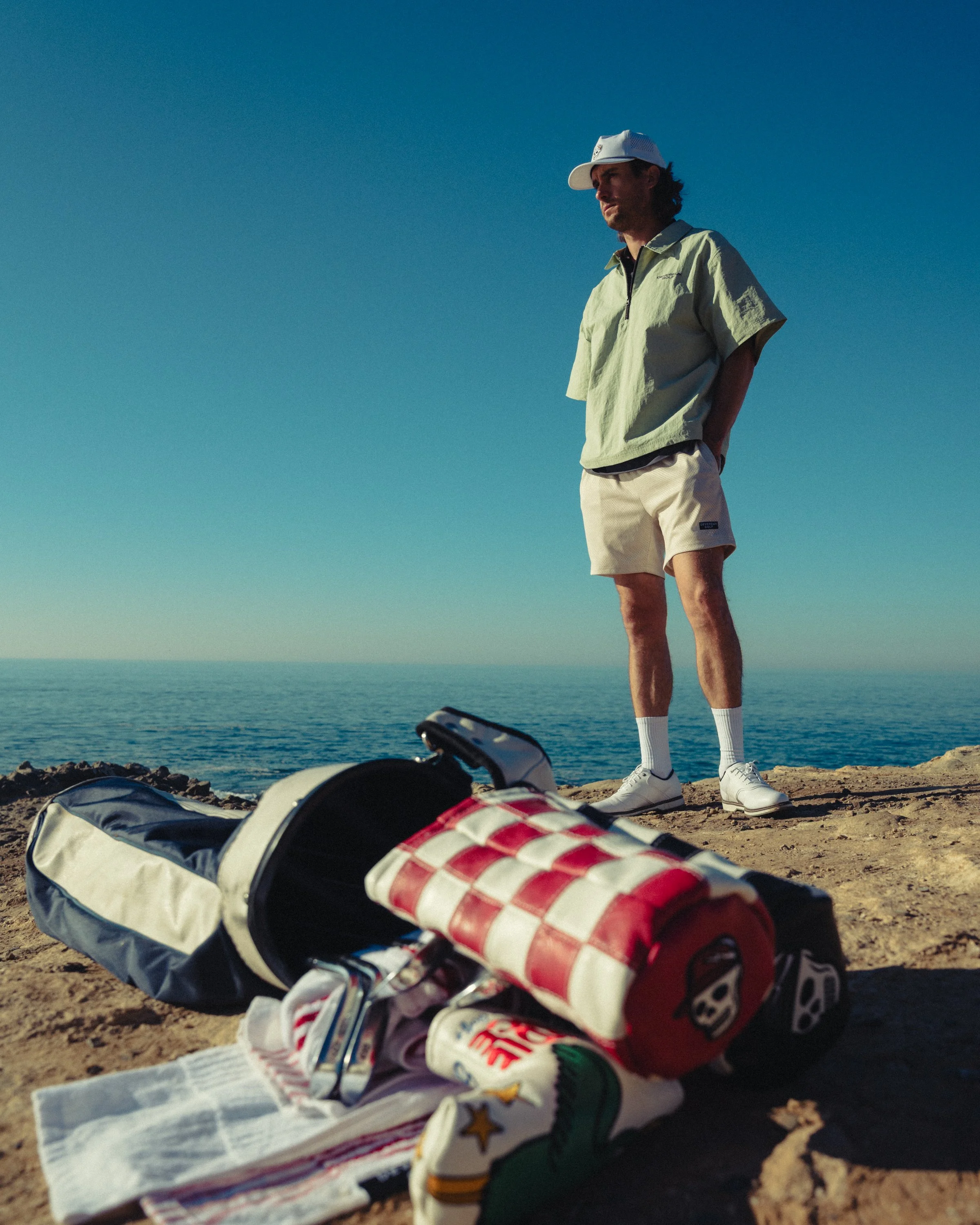 A man standing on a rocky cliff by the ocean, wearing a green jacket, cream shorts, white sneakers, and a white cap, with golf equipment and supplies in the foreground.
