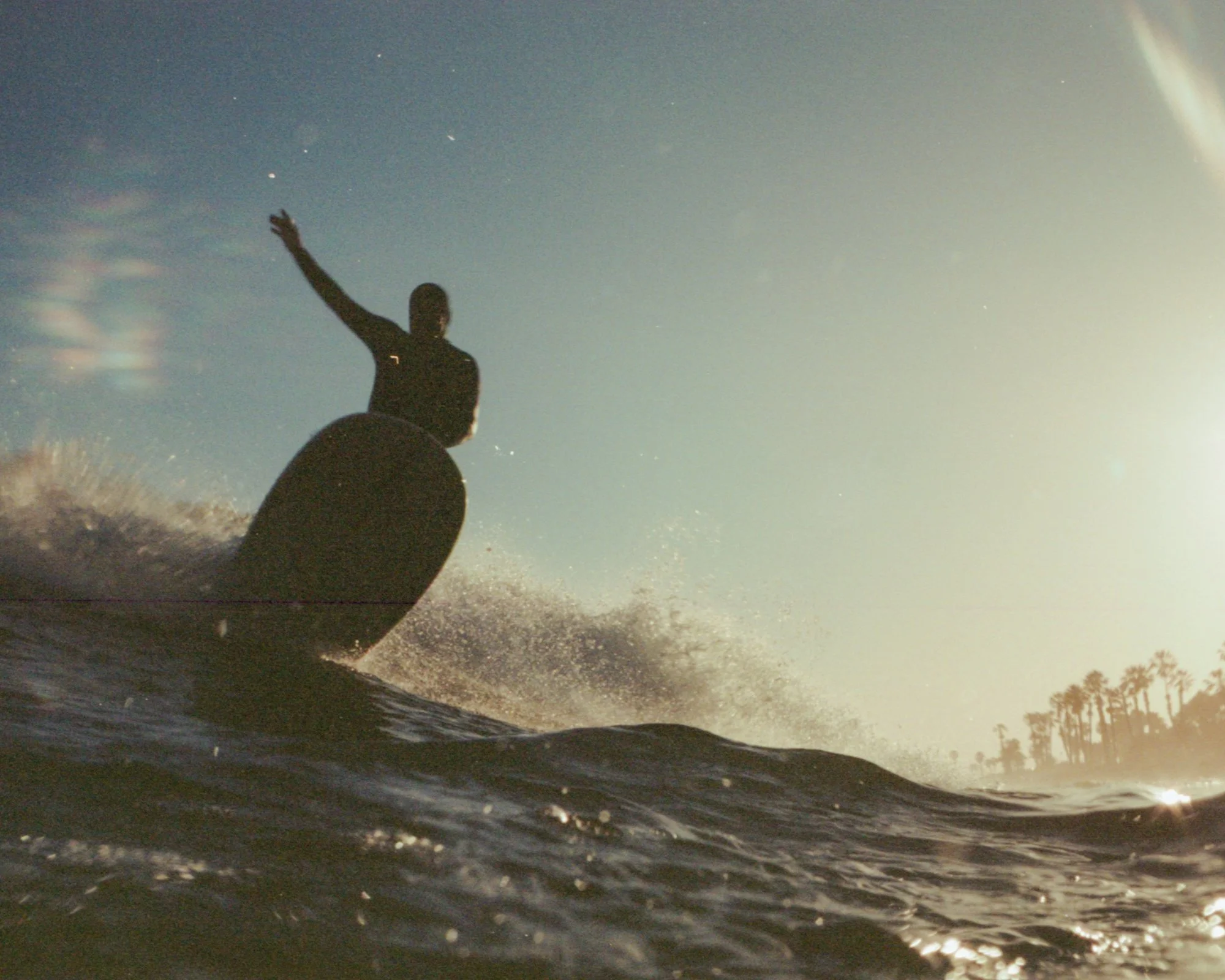 A silhouette of a person standing on a surfboard or boat, reaching out with one arm, against a cloudy sky with the sun shining, turbulent water, and distant trees on the shoreline.