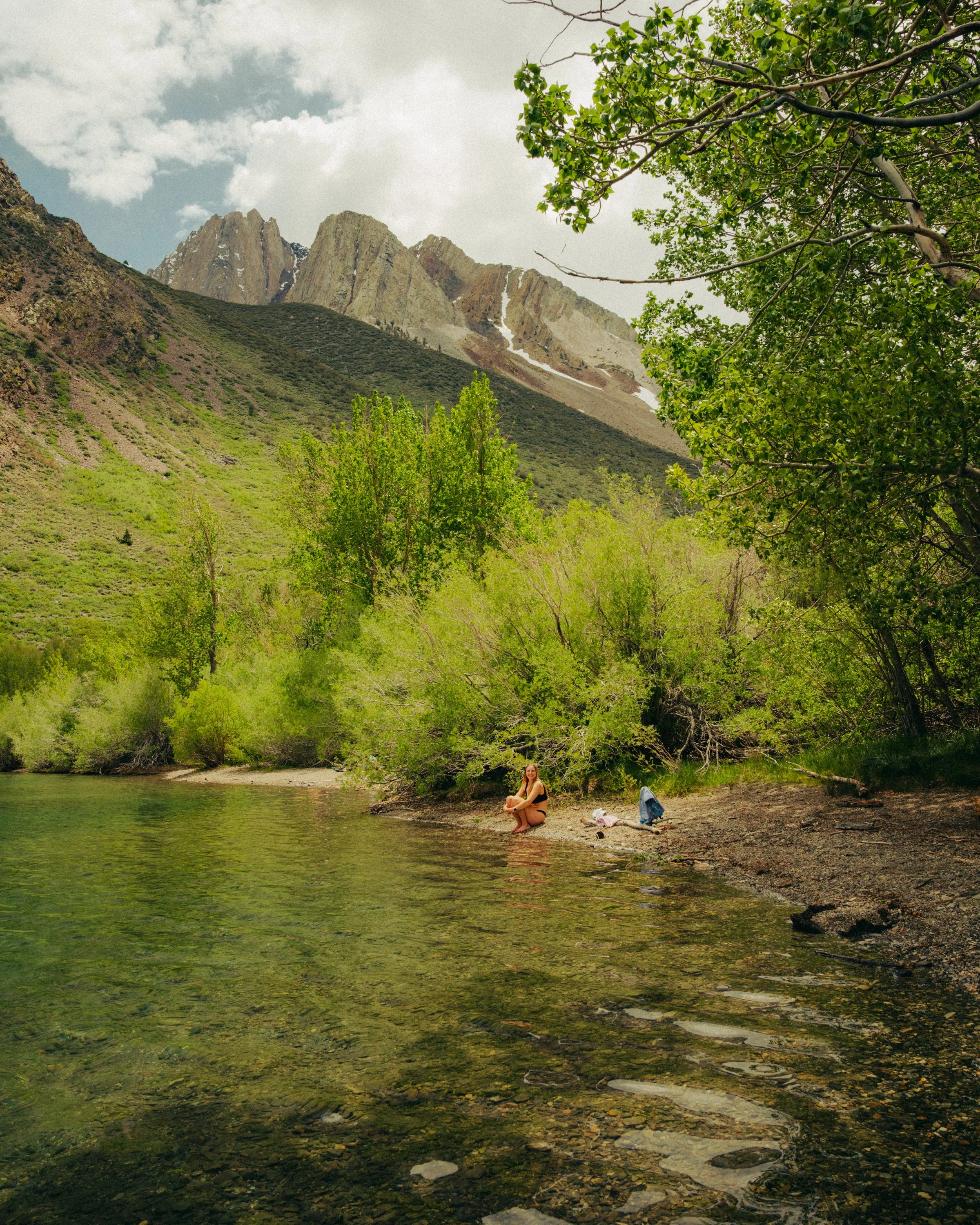 A woman sitting on the rocky shore of a river surrounded by lush trees with a mountain range in the background.