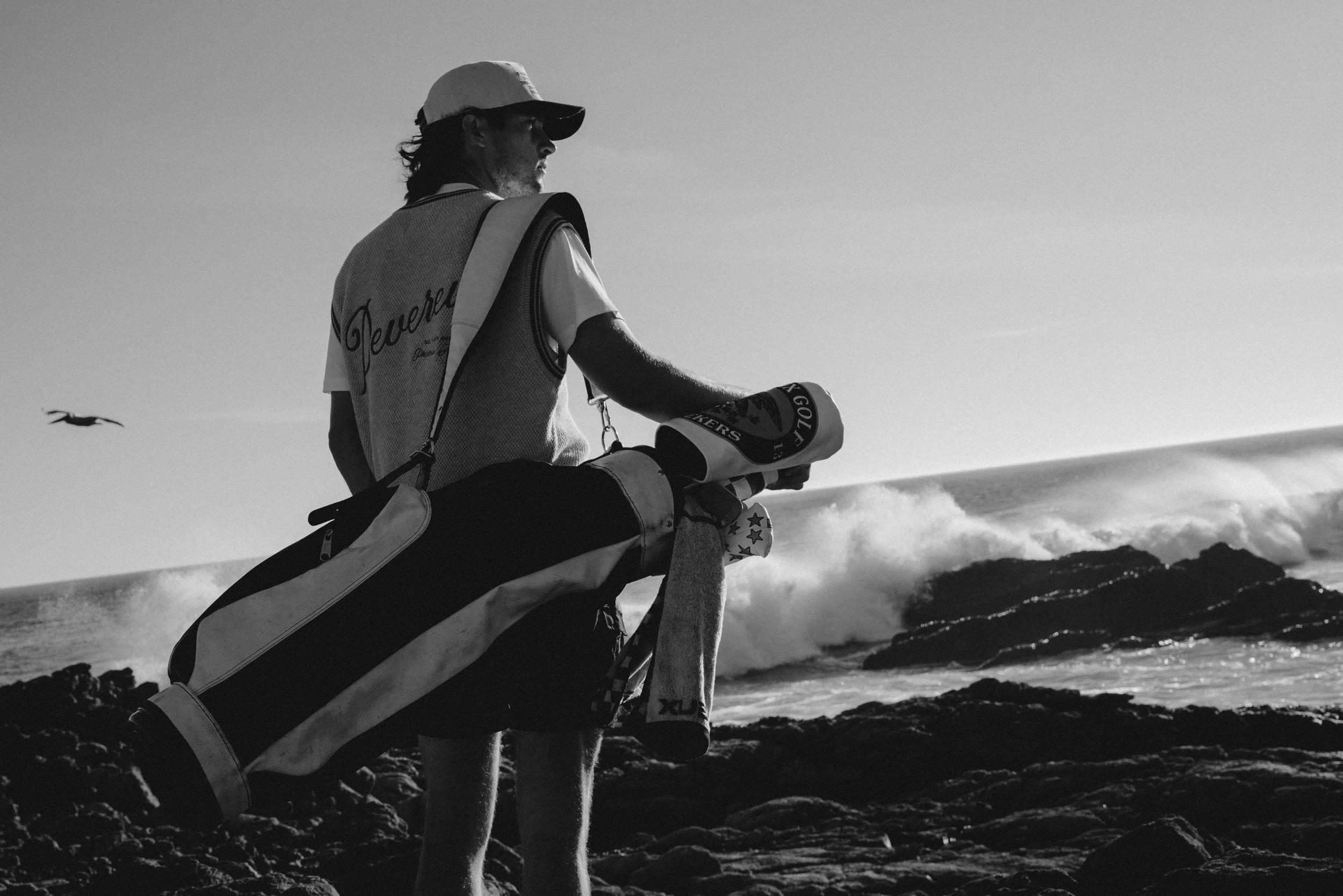 A man with a golf bag on his shoulder stands on rocks near the ocean, watching the waves roll in.
