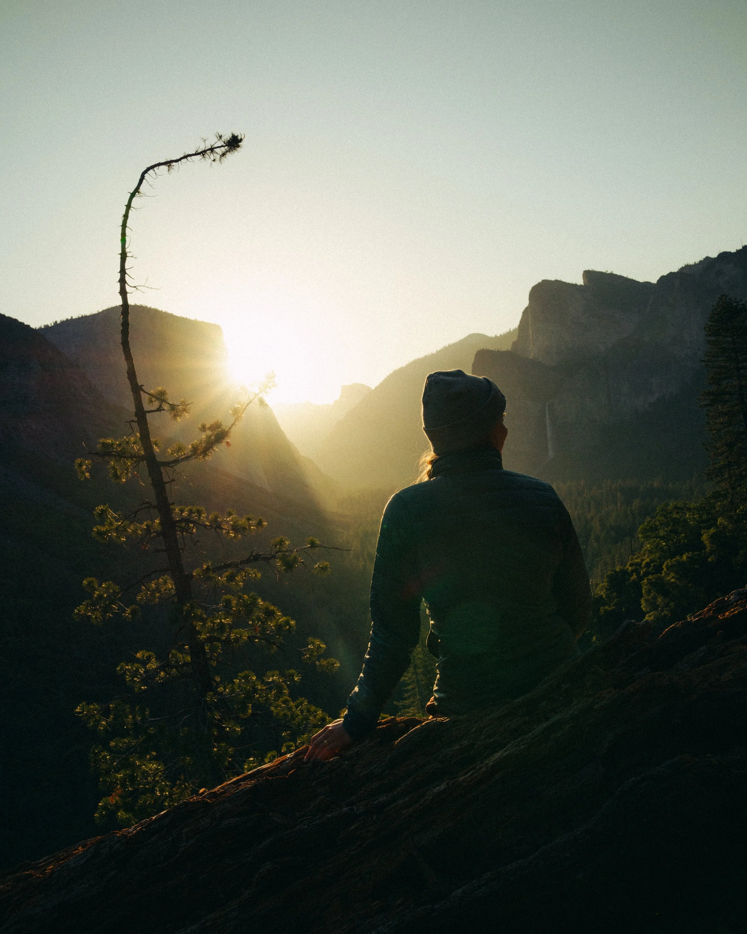 Person with dark hair and hat sitting on a rock, facing mountains and setting sun in a valley.