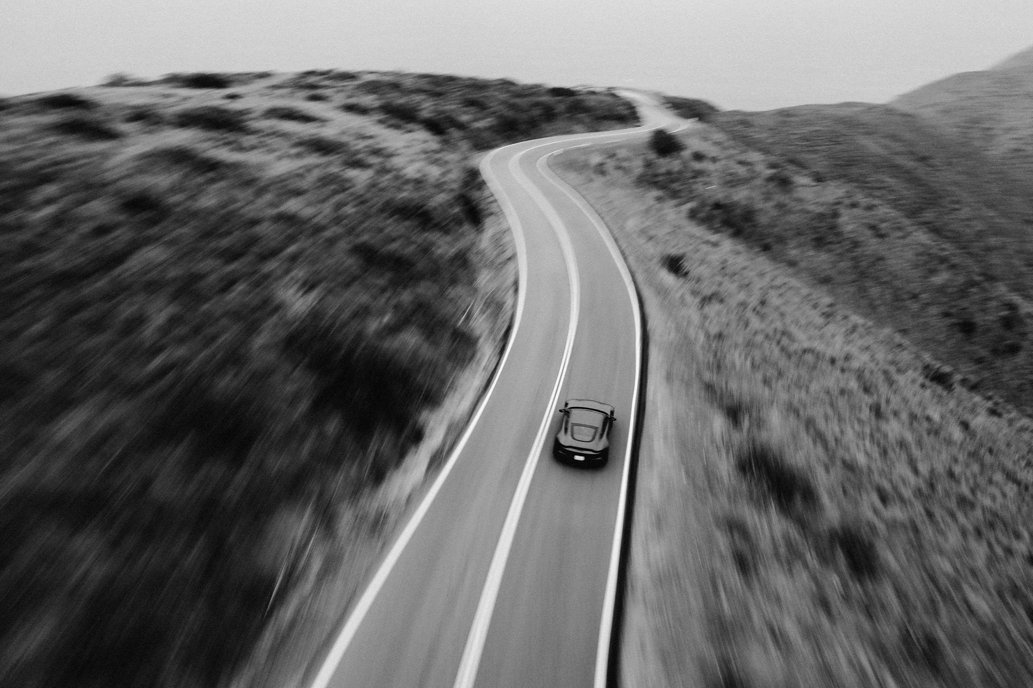 A black and white aerial view of a winding mountain road with a car traveling along it, surrounded by hilly terrain and sparse vegetation.