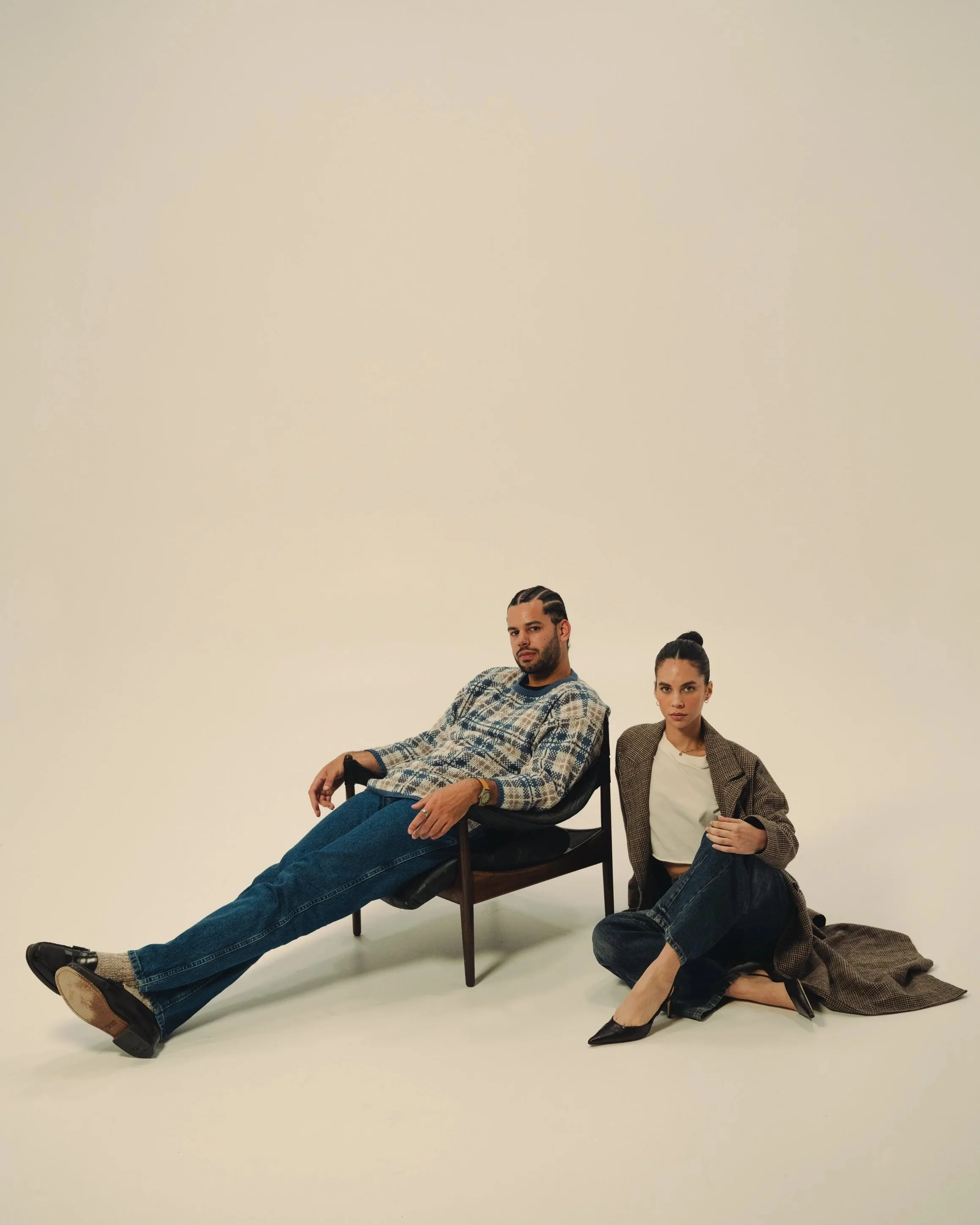 A man with dark hair and a woman with dark hair and light skin posing together in a minimalist studio with a beige background. The man is sitting in a chair with crossed legs, wearing a patterned shirt, jeans, and boots. The woman is kneeling and lea