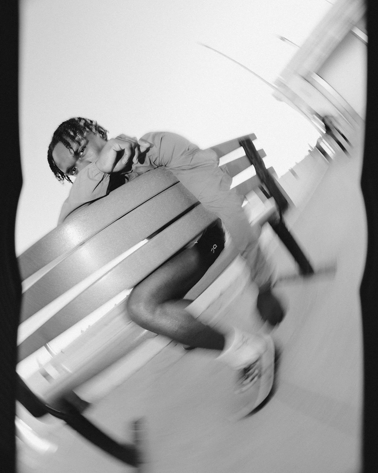 A black and white photo of a man sitting on a bench, with one leg crossed over the other, wearing a jacket, shorts, tights, and sneakers, looking directly at the camera, while resting his chin on his hand.