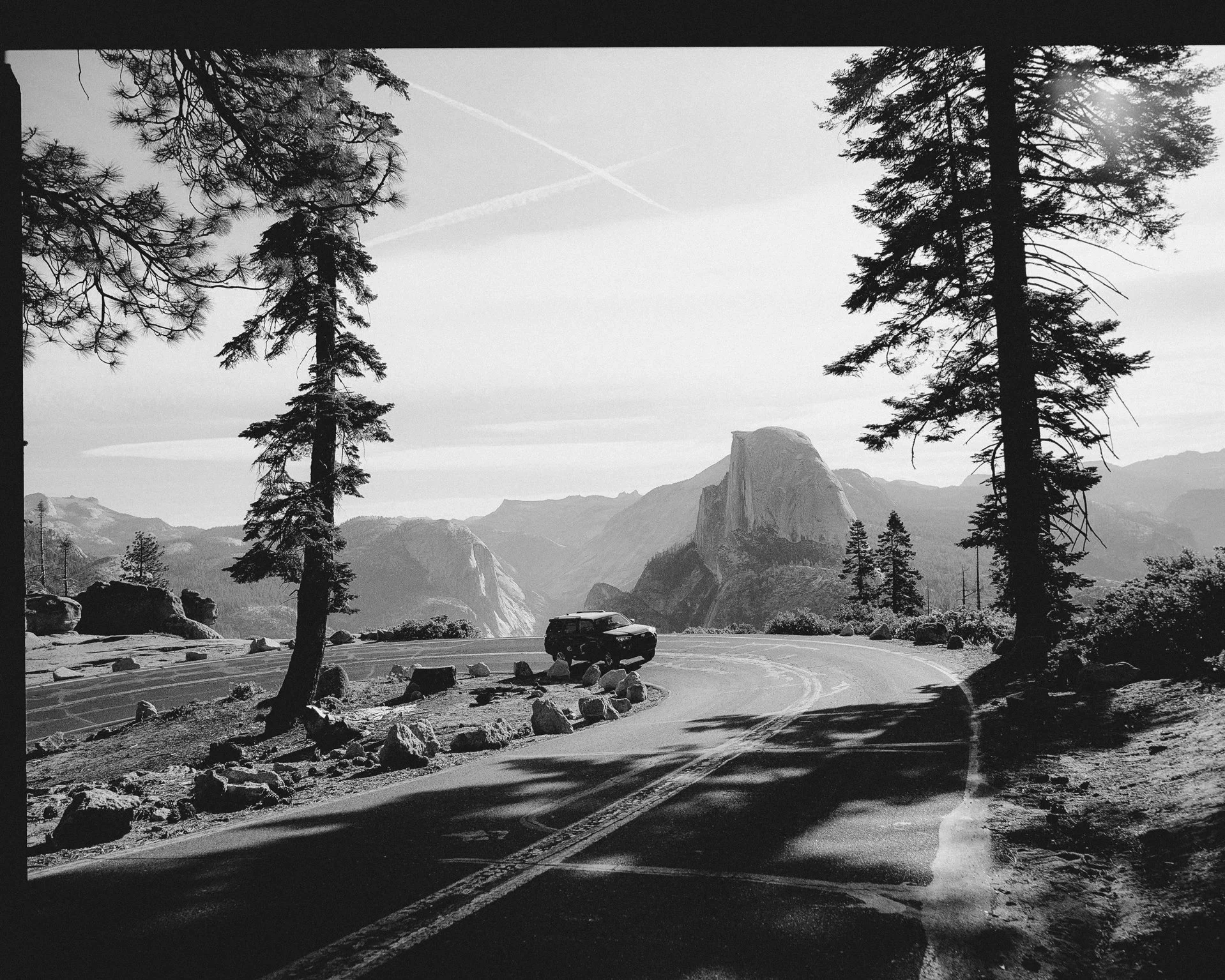 Black and white photo of a mountain landscape with tall trees in the foreground, a winding road, and large rock formations with mountains in the background.