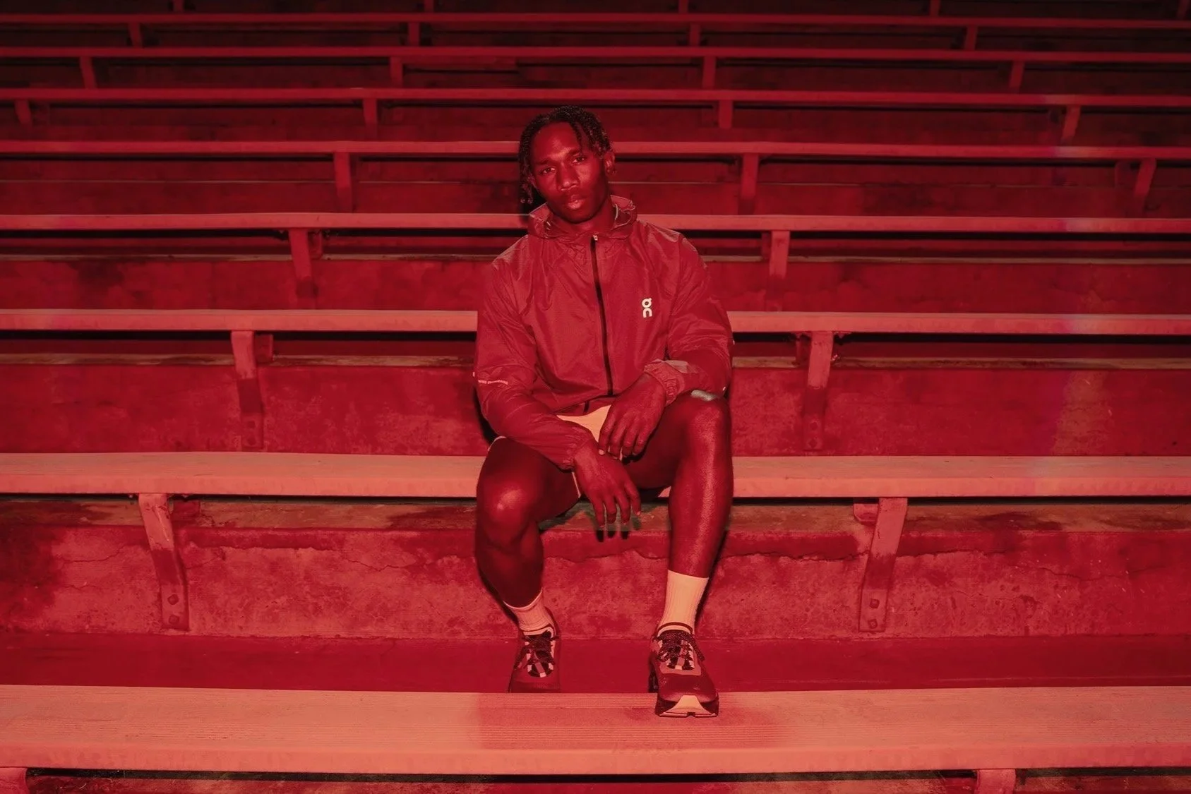 A young man sitting on wooden stadium bleachers in a red lighting environment, wearing a red jacket, black shorts, and sneakers.