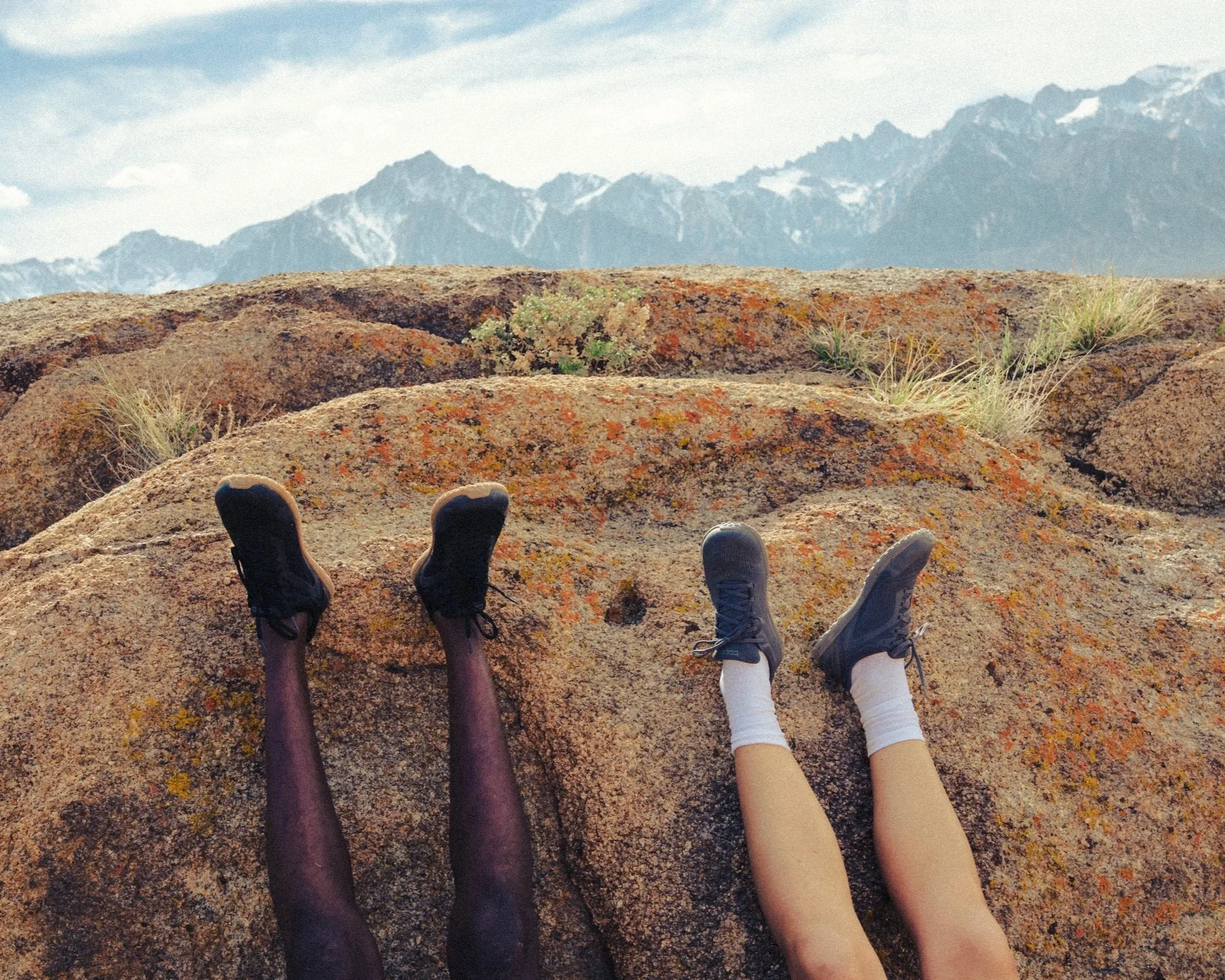 Four people with different skin tones sitting and lying on a large flat rock in a desert landscape with mountains in the background, wearing hiking shoes and shorts.