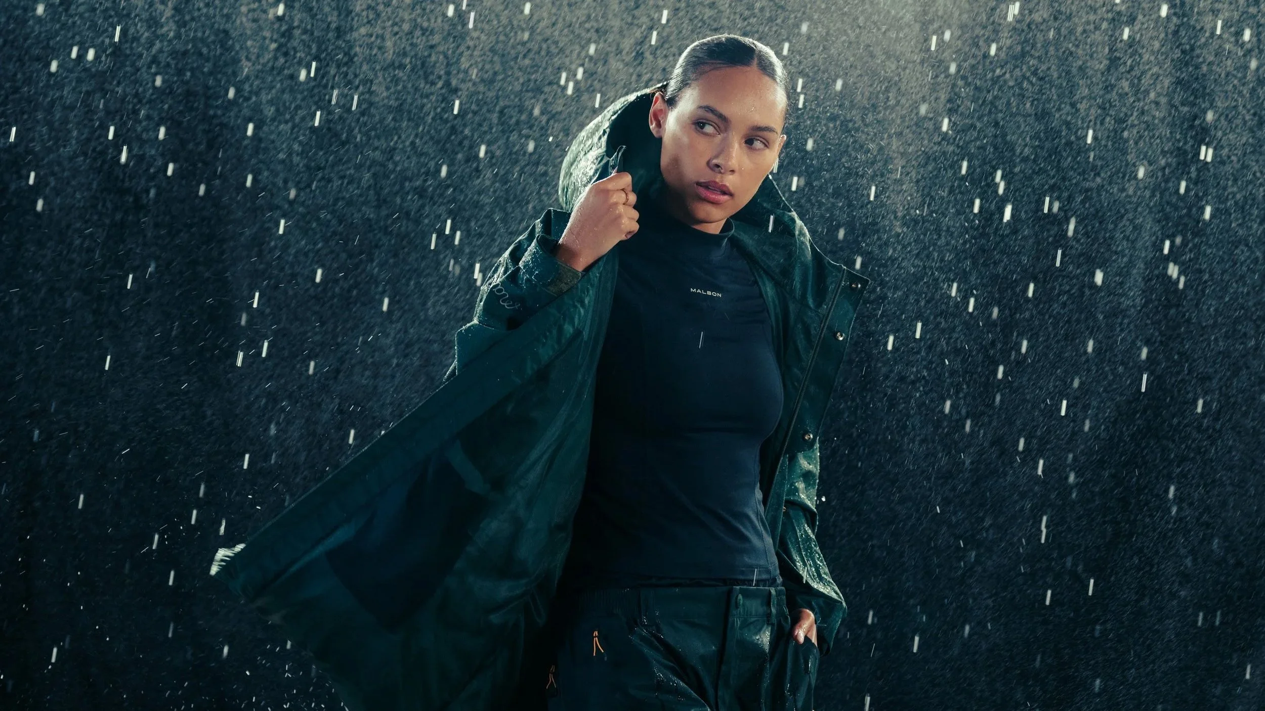 A woman with wet hair and a rain jacket standing in pouring rain, looking to her left.