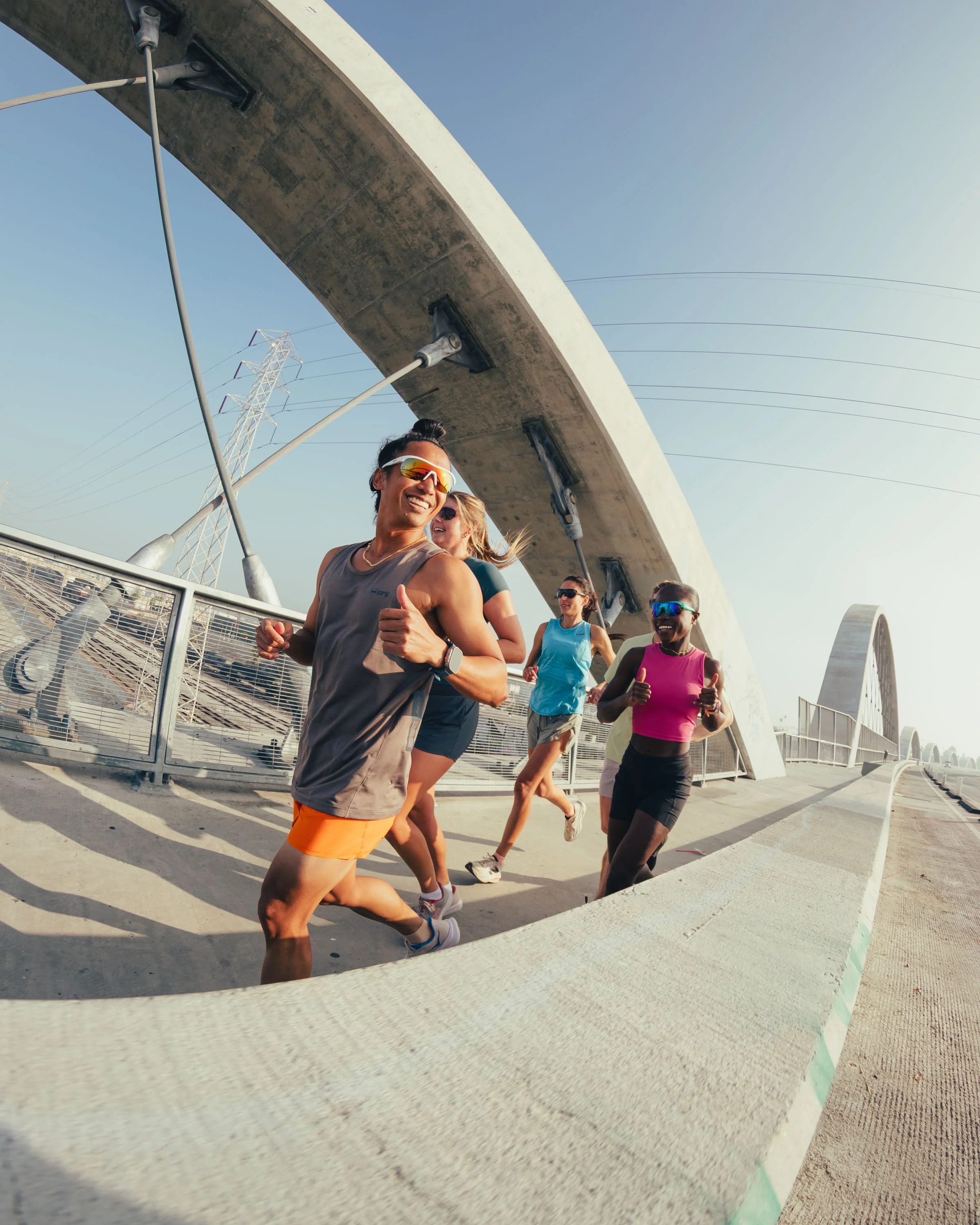 A group of five diverse runners jogging on a bridge under a clear sky, smiling and wearing athletic clothing and sunglasses.