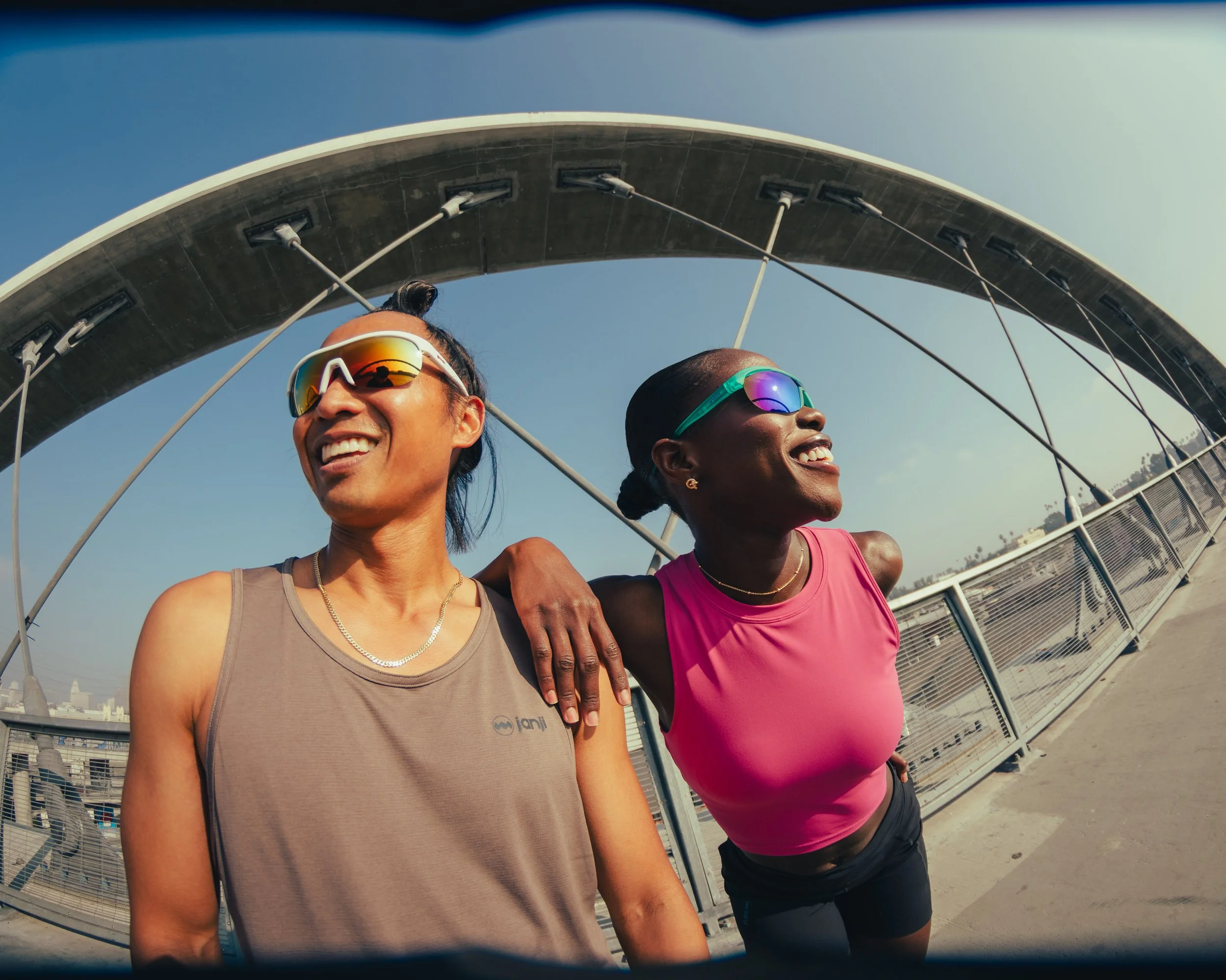 Two smiling women in athletic gear, wearing sunglasses, enjoying a sunny day outdoors under a bridge with a city skyline in the background.