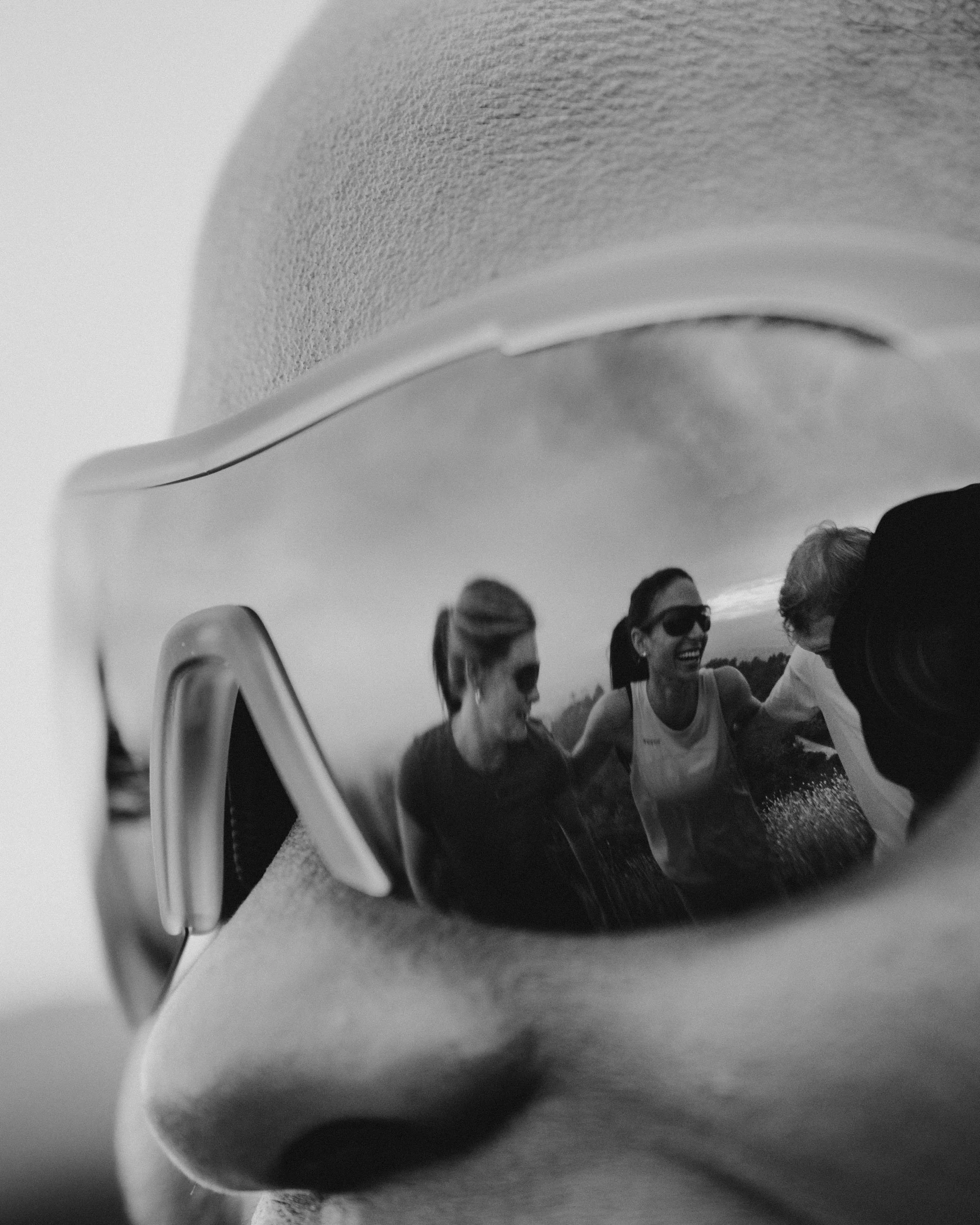 Close-up of a person's face wearing sunglasses, with a reflection of three women smiling and talking outdoors in the sunglasses' lenses.