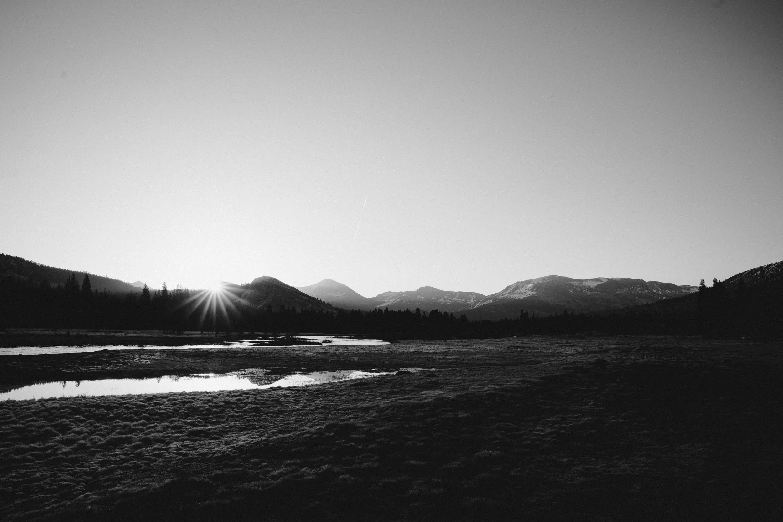 Black and white photo of a mountain landscape with a lake in the foreground and the sun setting behind the mountains.