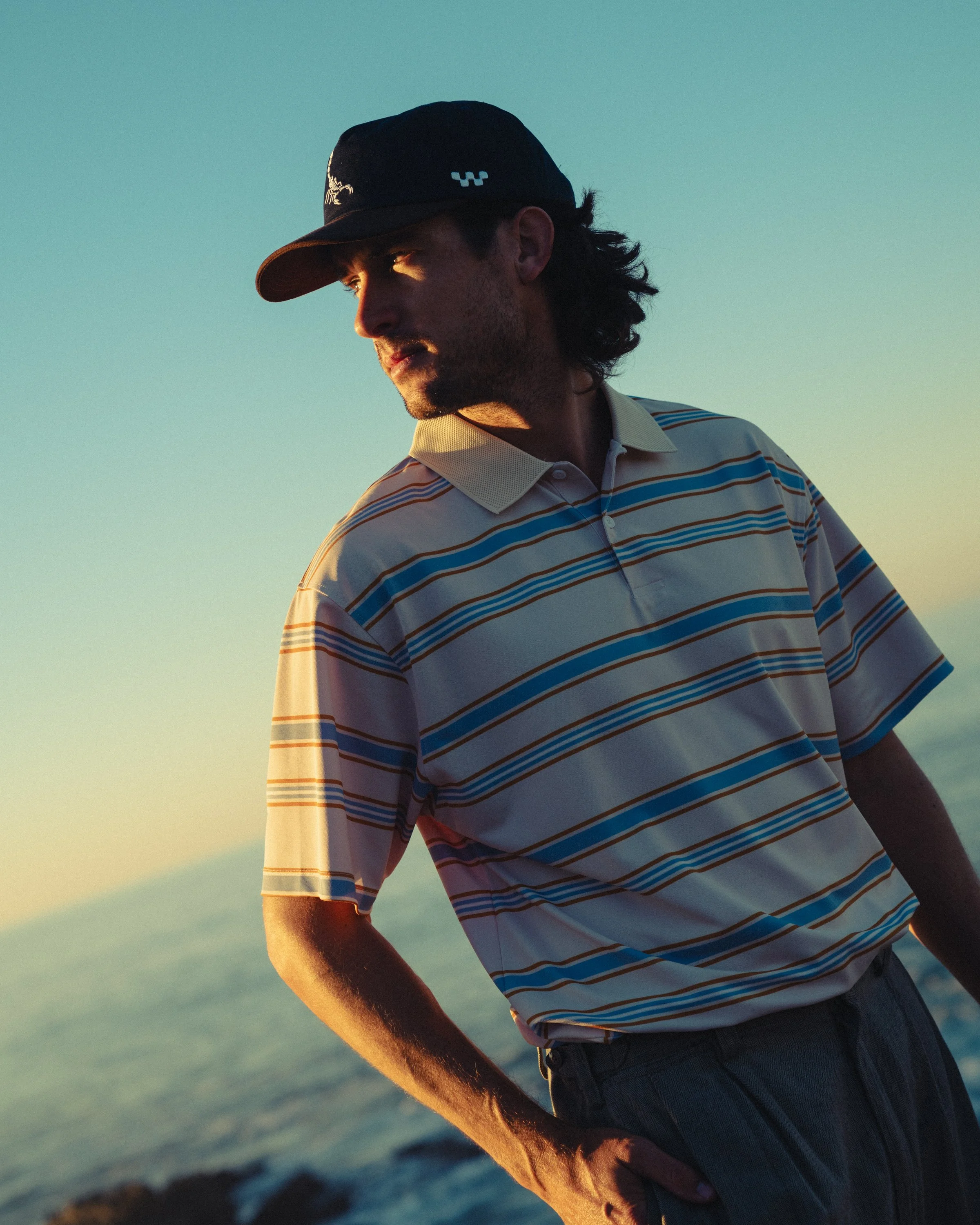 A man in a baseball cap and striped polo shirt stands near the ocean during sunset.