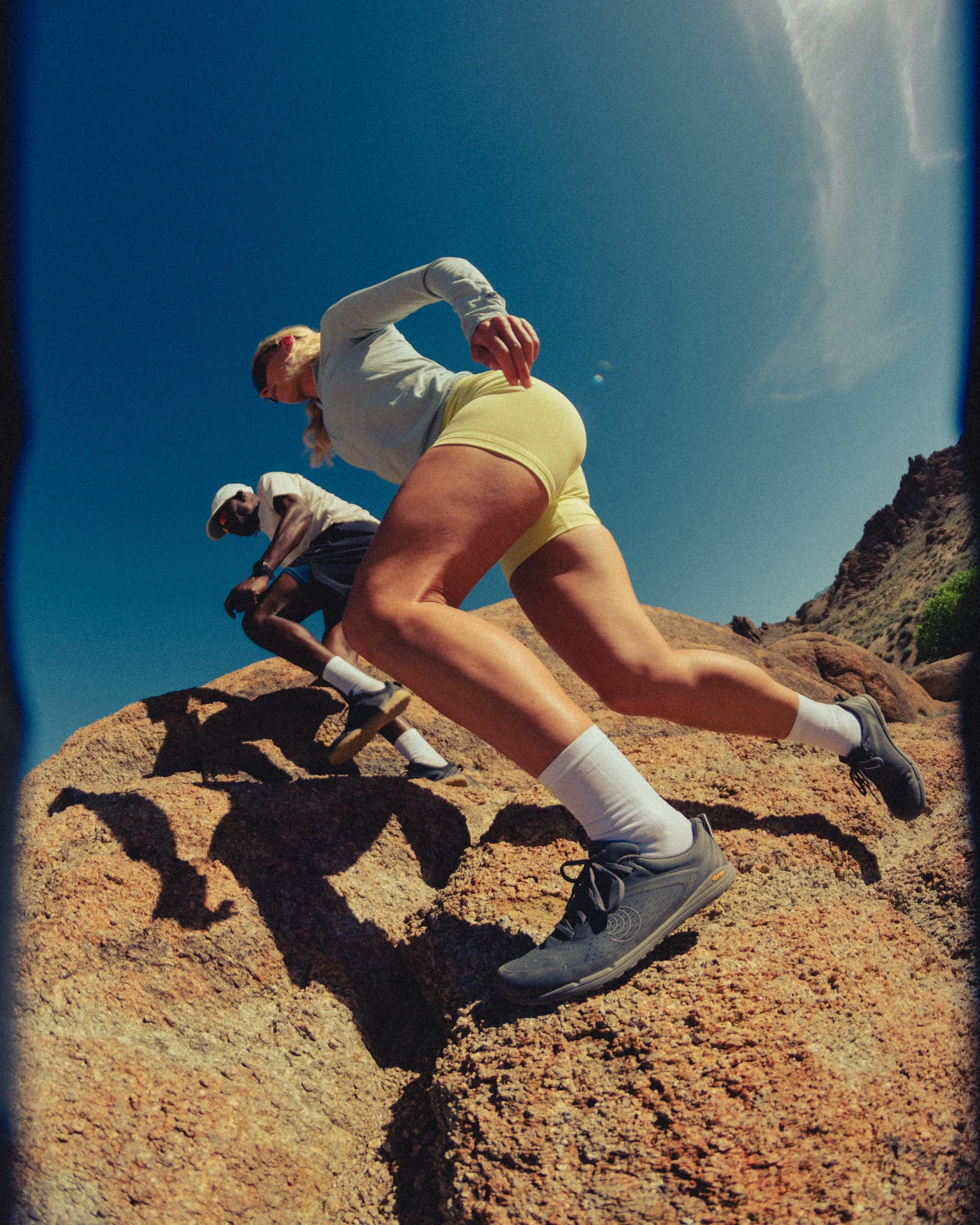 A woman in yellow shorts and a white hoodie climbing a rocky hill, shadow cast on the ground, clear blue sky, other people in the background, sun shining, desert landscape.