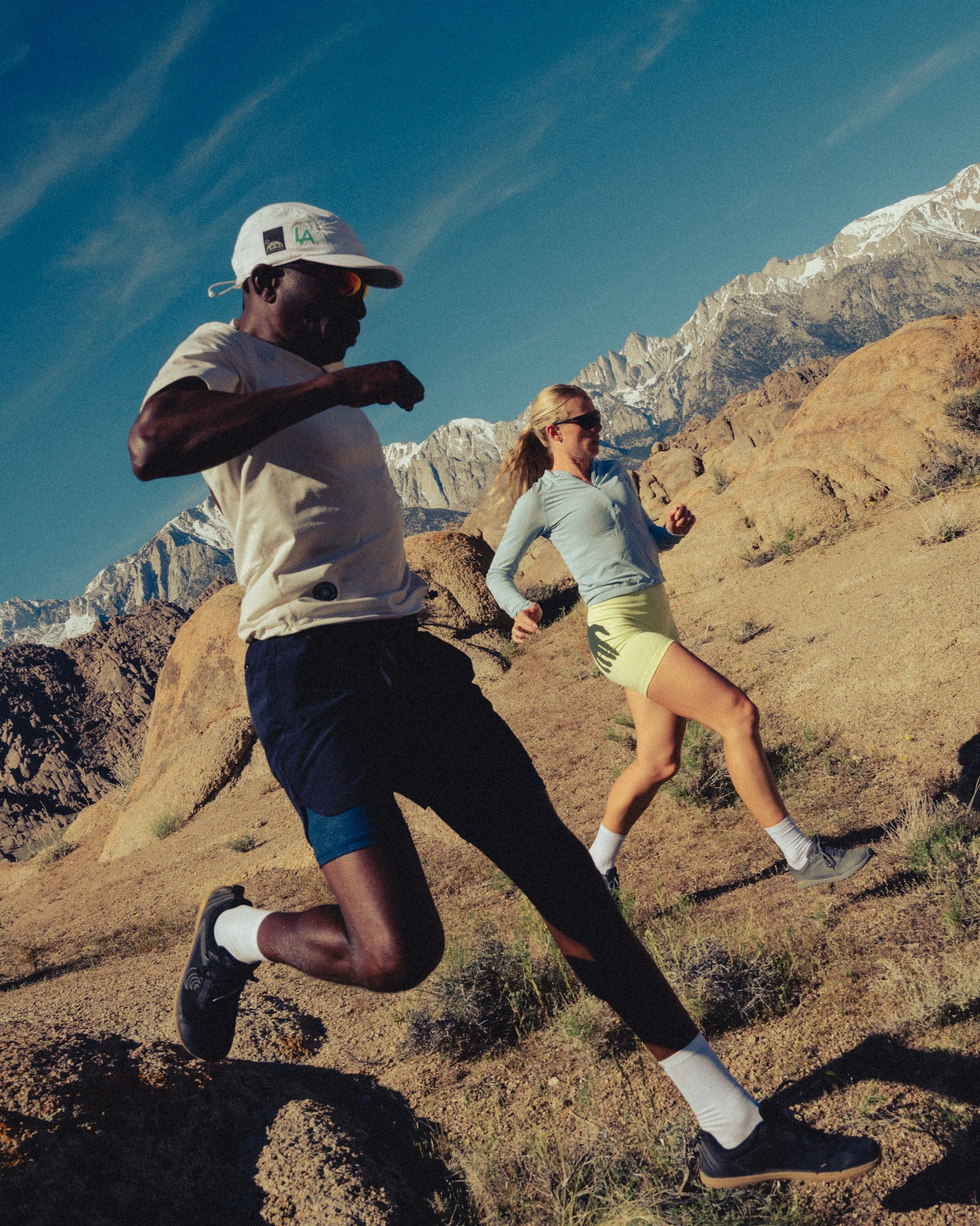 Two runners, a man and a woman, jogging on a rocky trail in a mountainous area with snow-capped peaks in the background on a clear day.