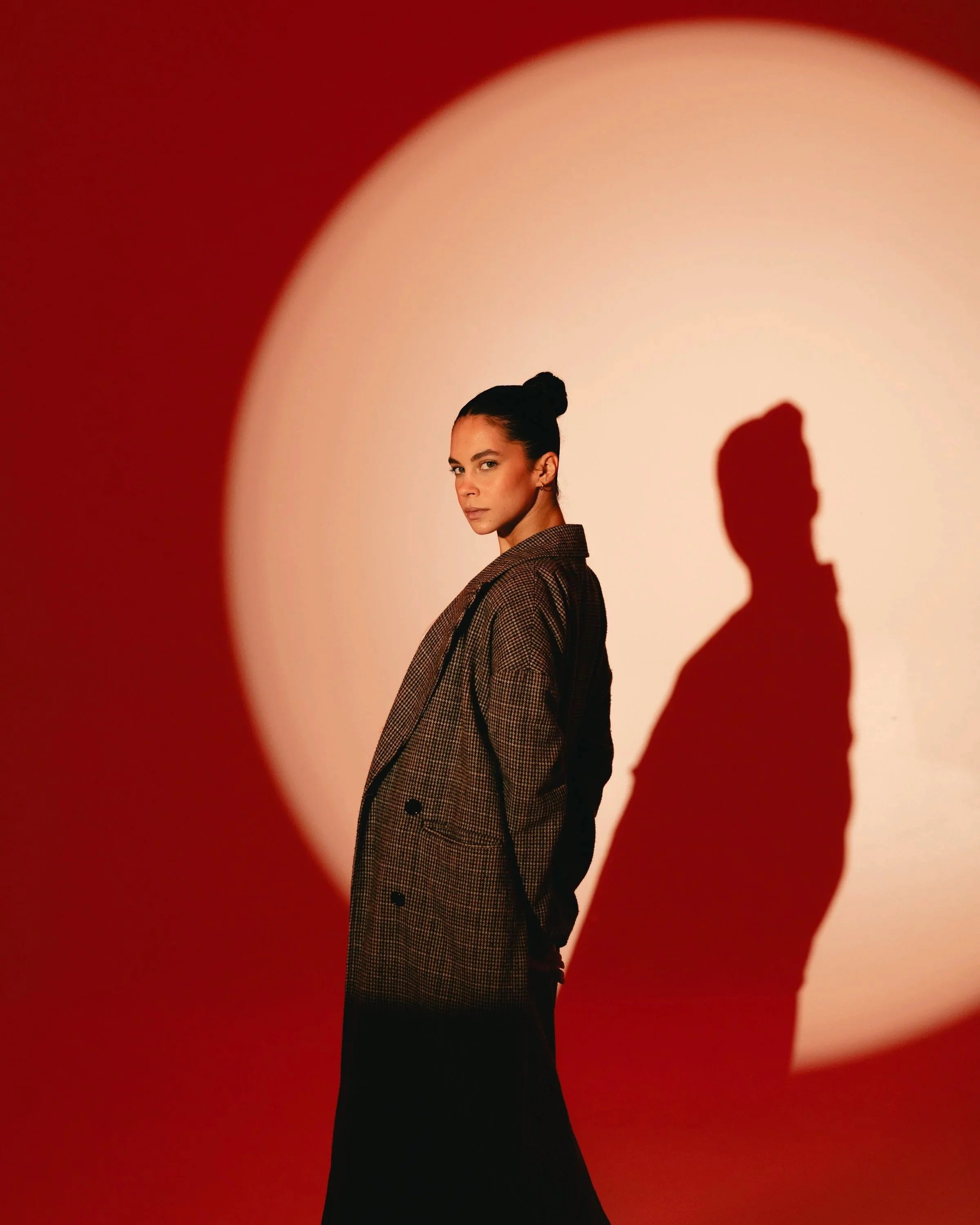 A woman with dark hair tied in a bun, wearing a brown plaid blazer, stands with her back to the camera but turns her head to look at the camera. Her shadow is cast on a wall with a bright, circular light behind her, creating a dramatic effect.