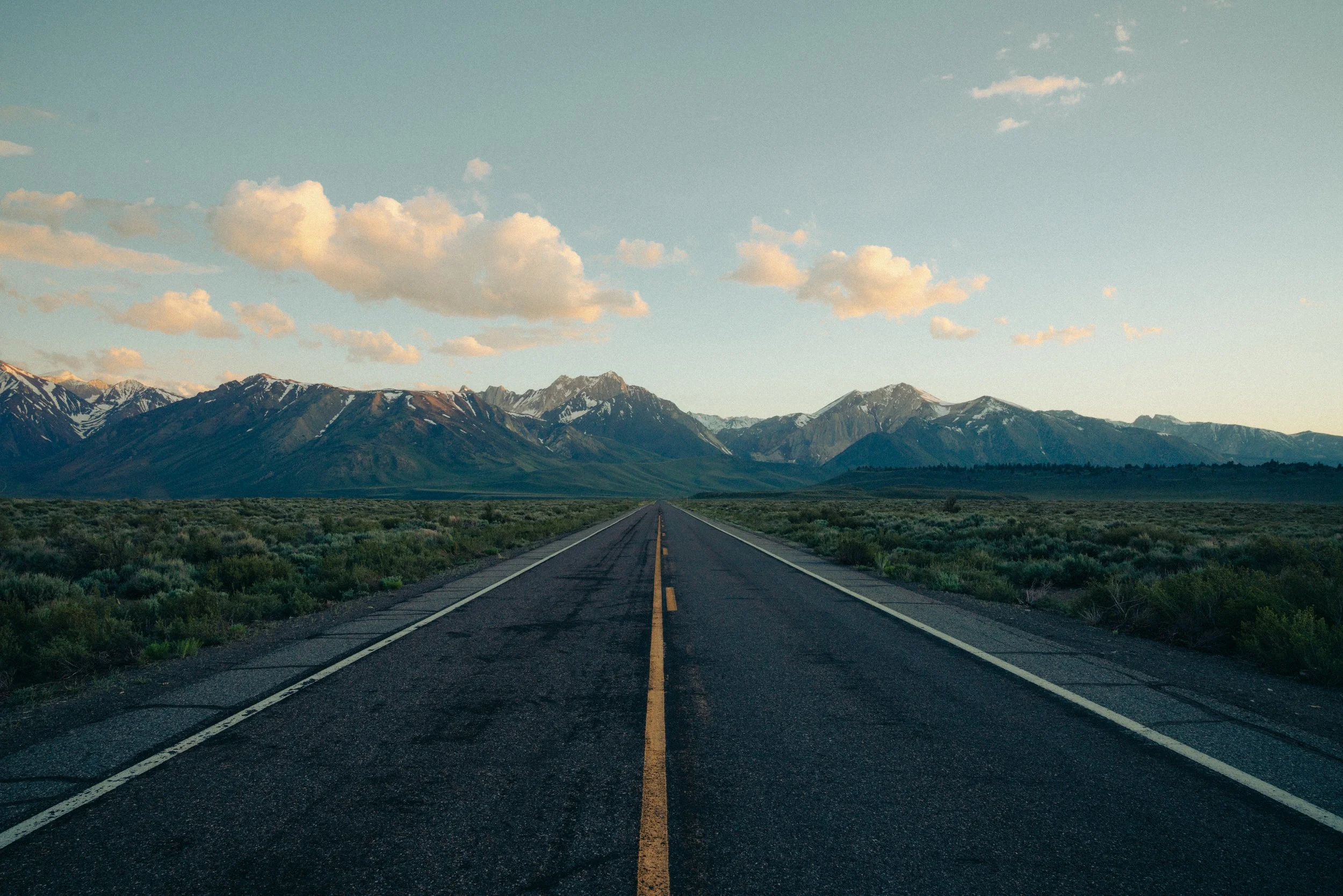 Long empty road leading toward snow-capped mountains under a partly cloudy sky.