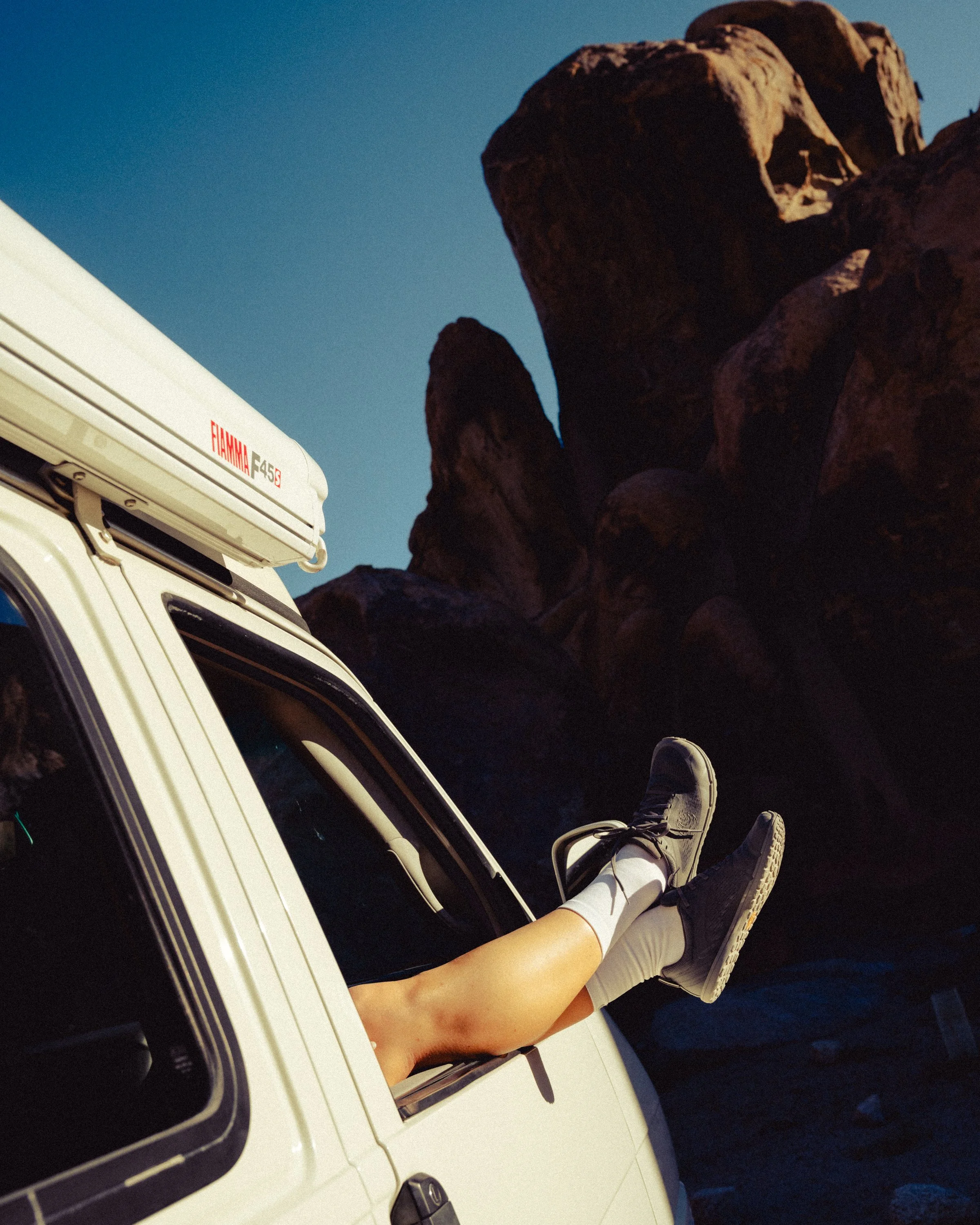 Photo of a person in shorts and sneakers lying inside a white vehicle with their legs hanging out of the window, against a backdrop of dark rocky formations and a clear blue sky.