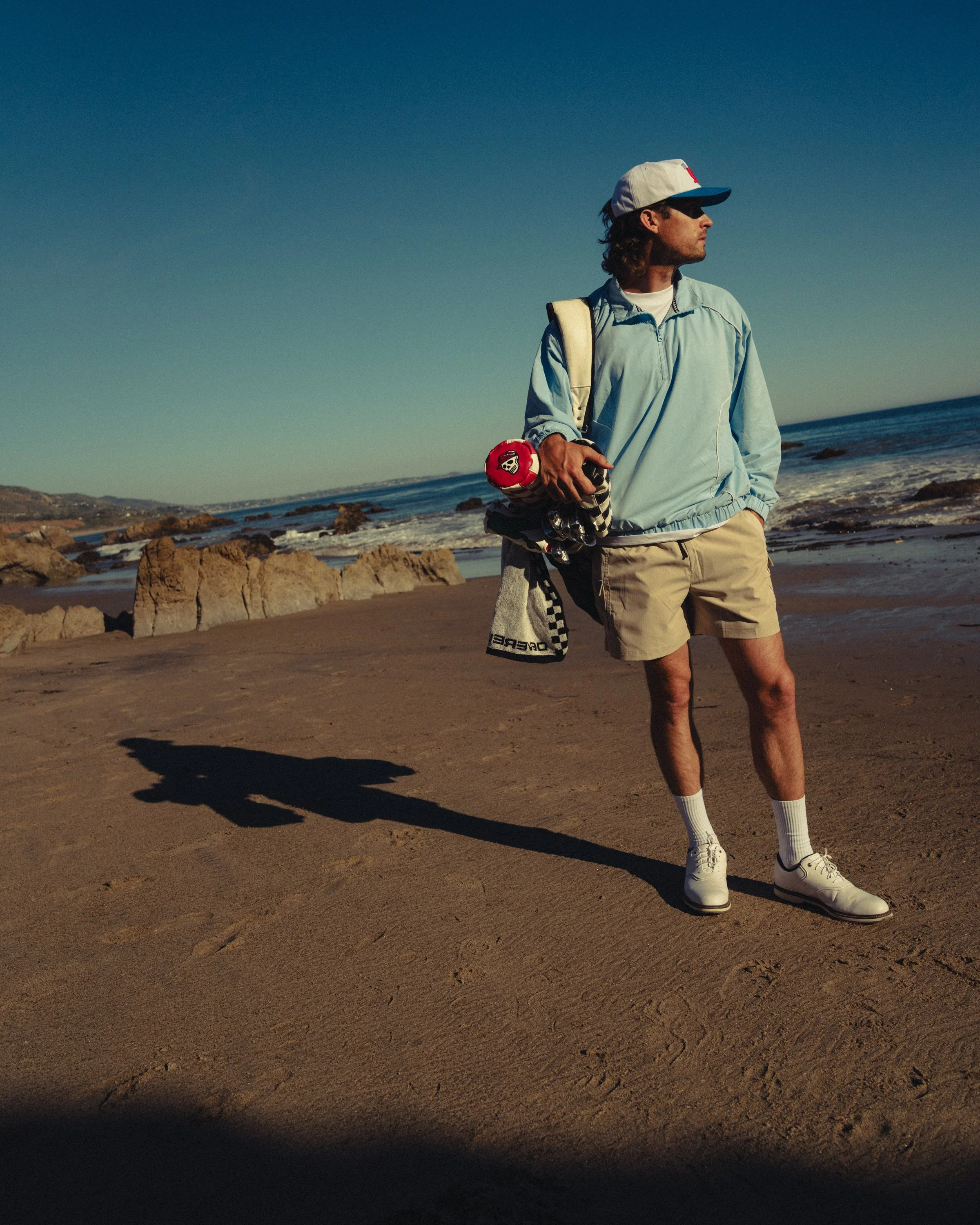 A man standing on a sandy beach holding a golf club and a golf ball, dressed in casual golf attire, with ocean and rocks in the background.