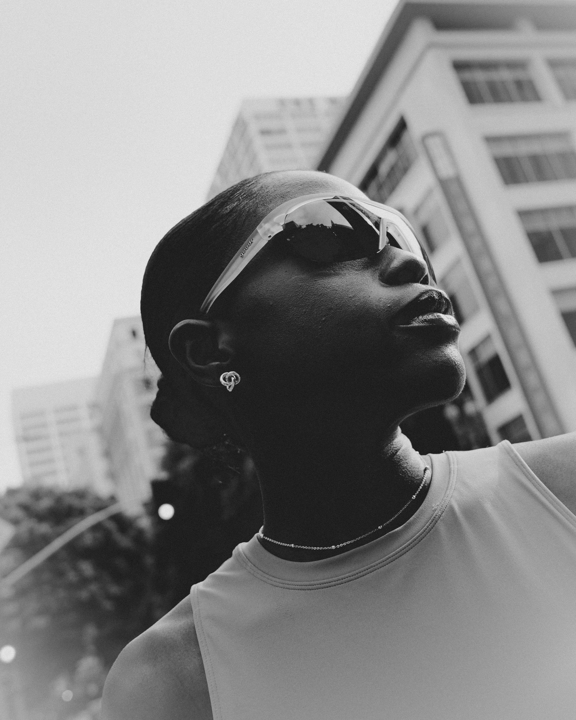 Black and white photo of a woman with sunglasses, earrings, and a necklace, outdoors in an urban area, with buildings in the background.