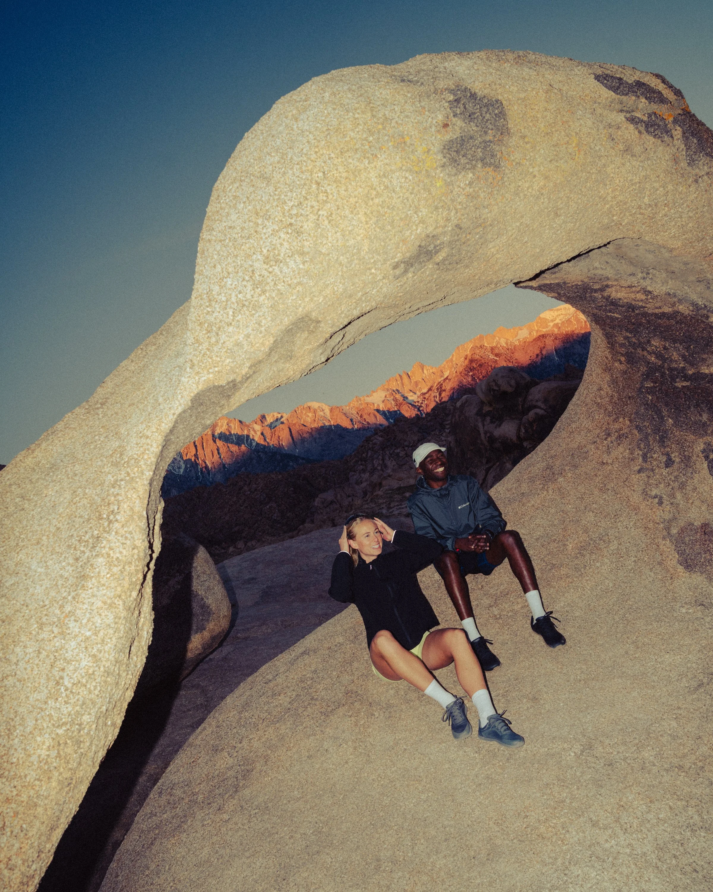 Two smiling women sitting under a large natural rock arch formation at sunset, with mountain range in the background.