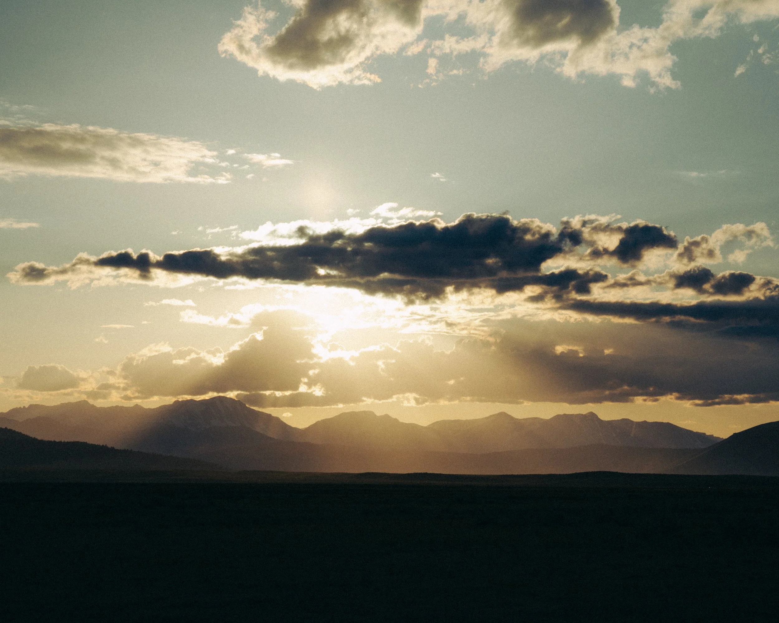 A landscape view of distant mountains under a partly cloudy sky with the sun setting or rising, casting a warm glow.