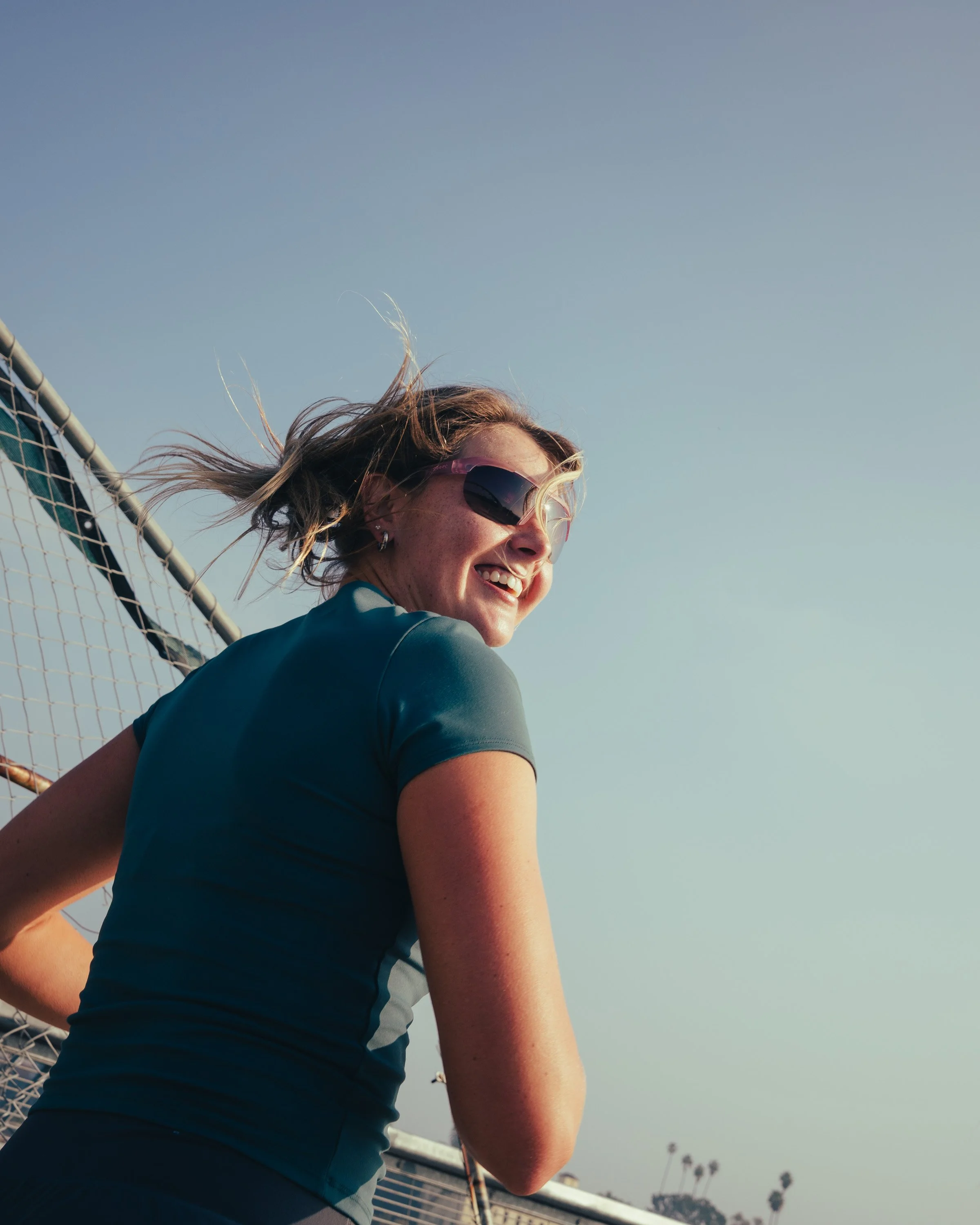 A woman with sunglasses smiling, wearing a sports top, on a running track during daytime.