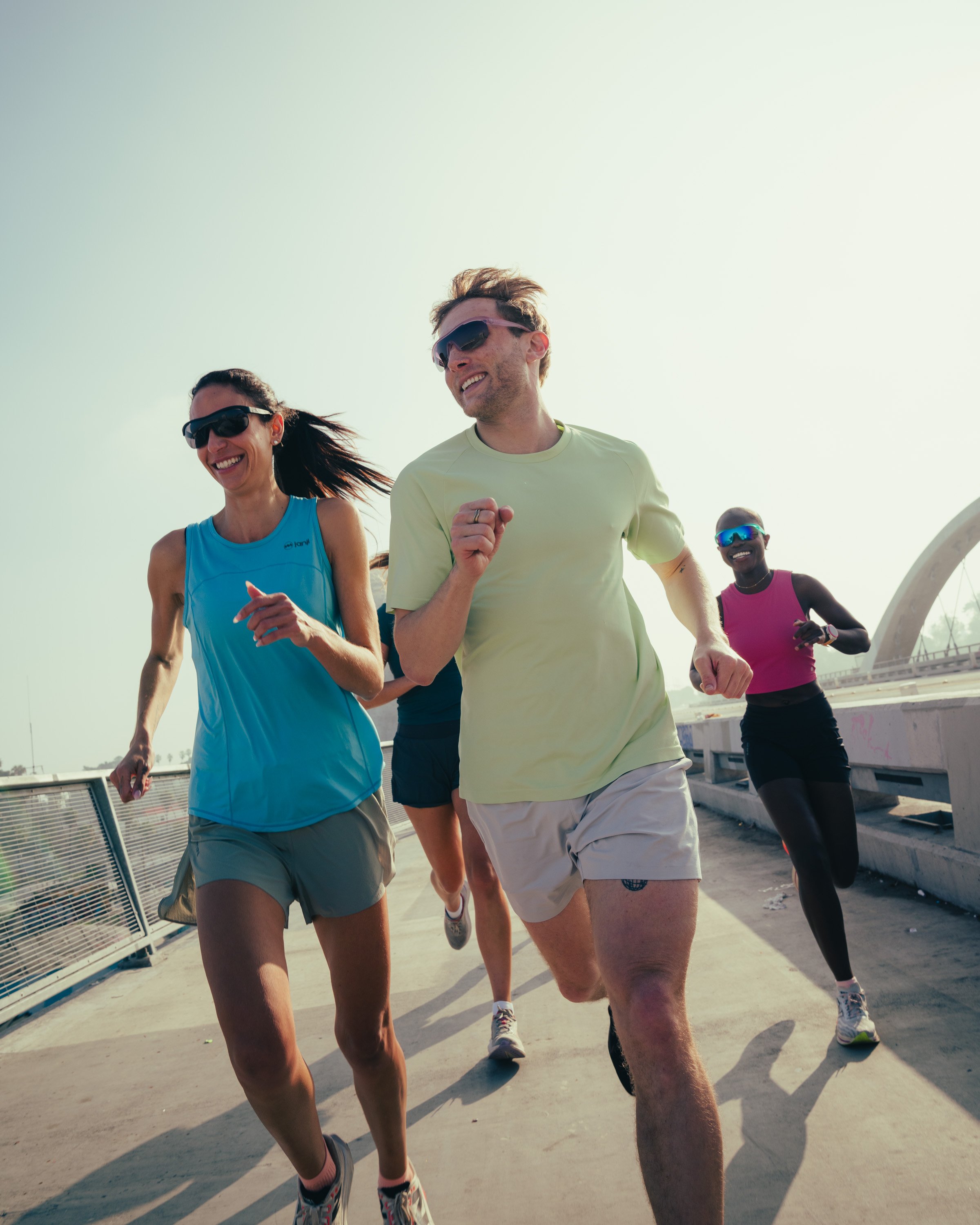 Group of people running outdoors on a bright day, wearing athletic clothing and sunglasses, smiling and enjoying exercise.