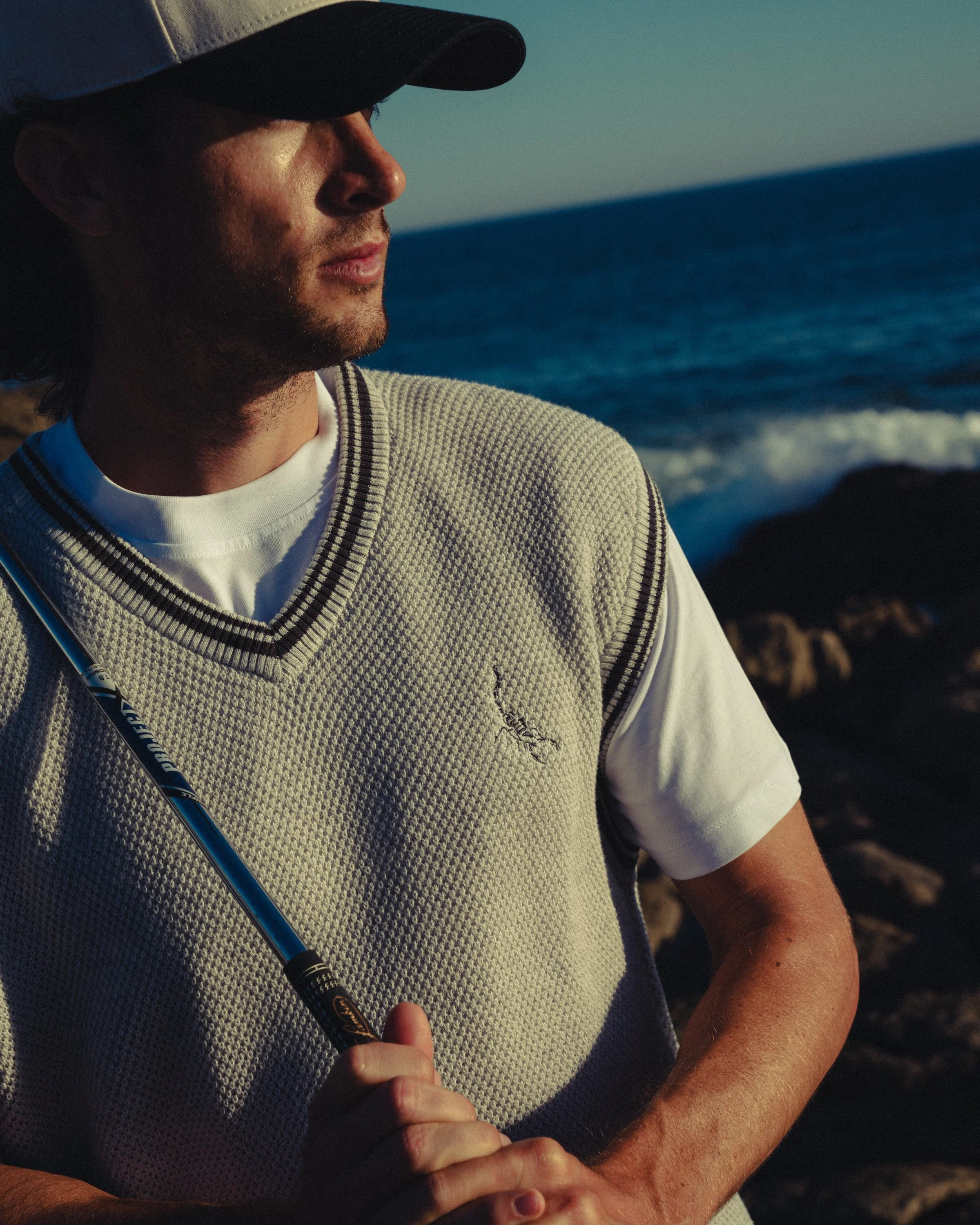 A man wearing a baseball cap, white shirt, and sleeveless sweater holding a golf club near the ocean during sunset.