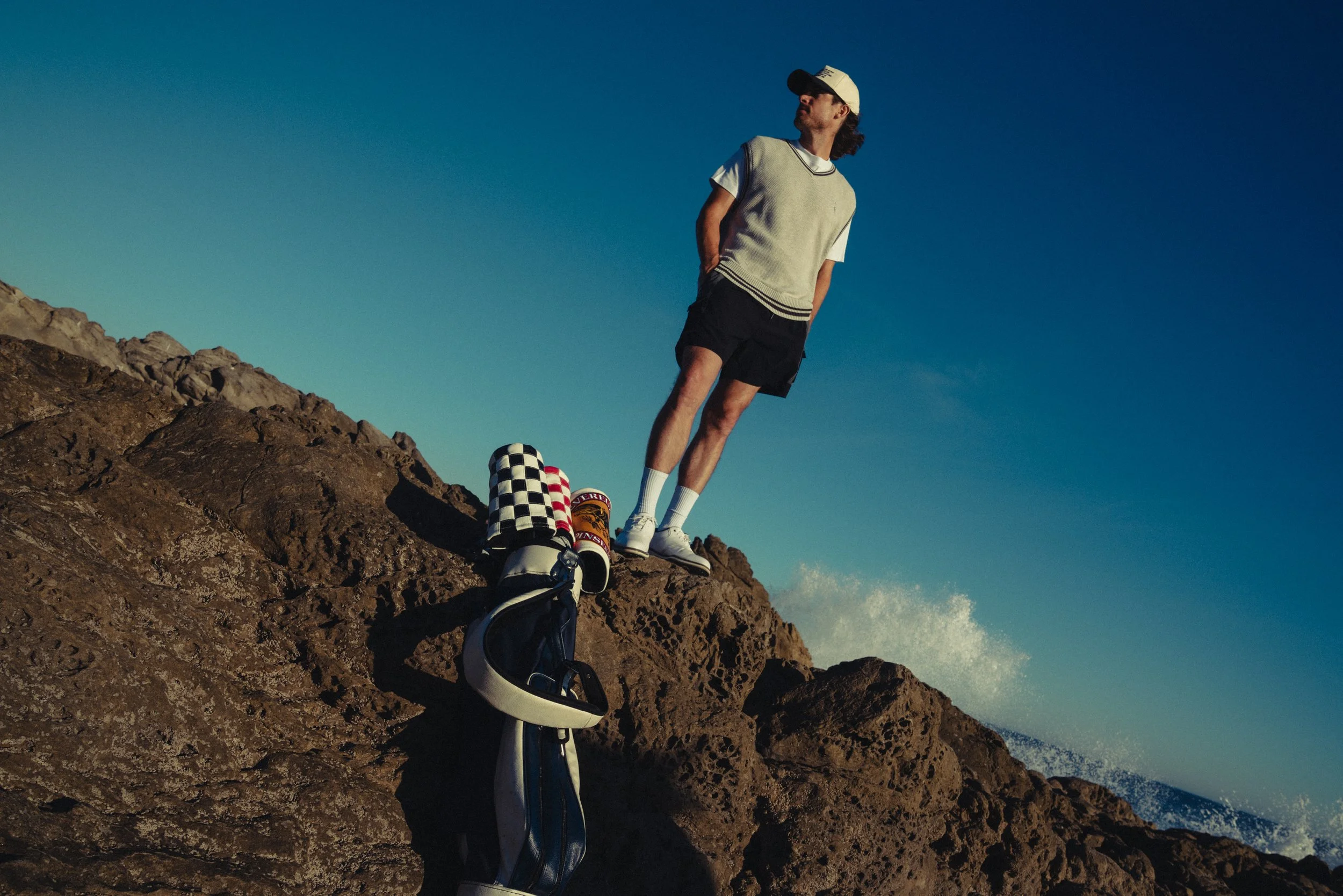 A man standing on a rocky cliff with a golf bag containing clubs, by the sea during sunset or sunrise.