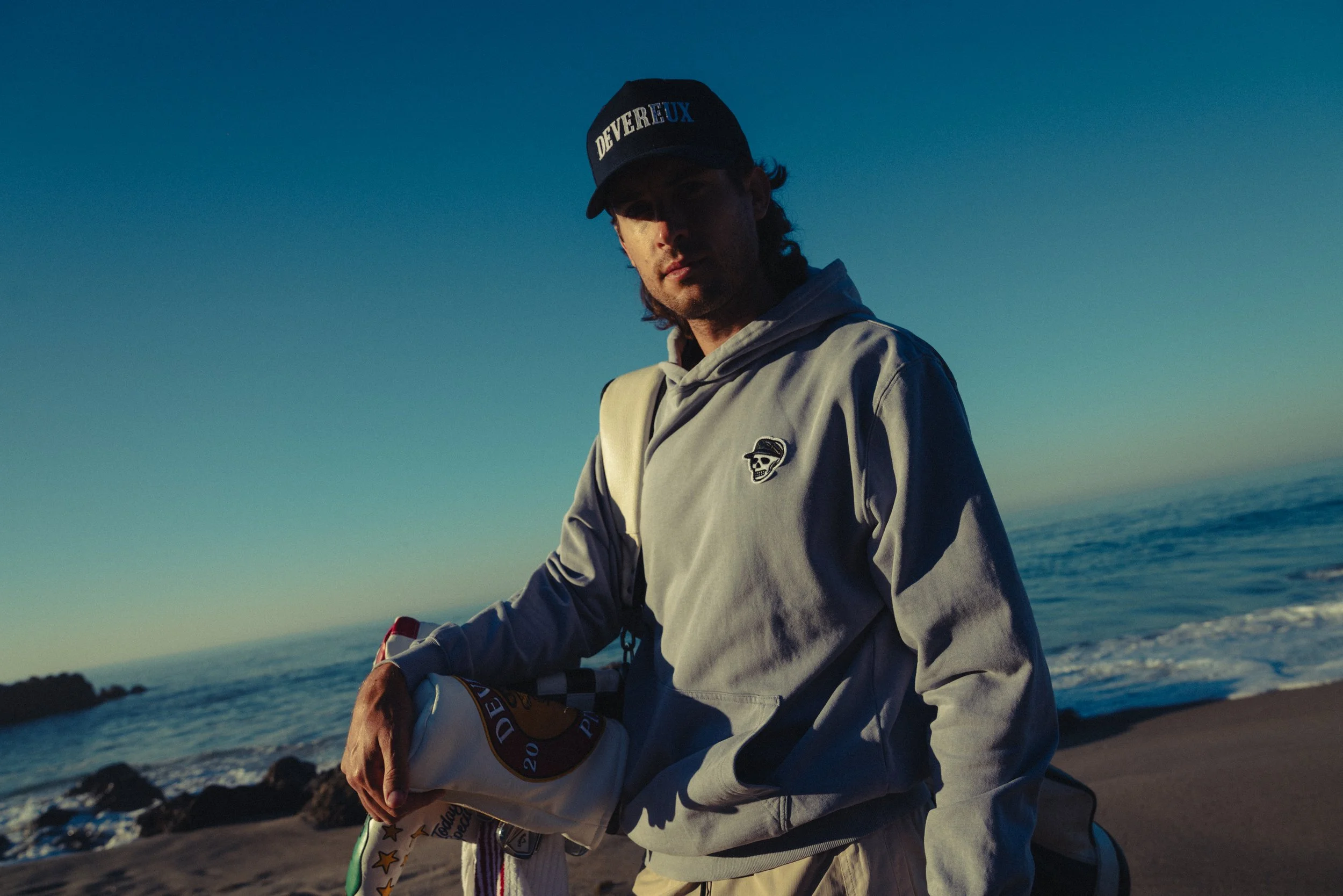 A young man at the beach, holding a surfboard, wearing a gray hoodie with a skull logo and a black cap that says 'Deuverux', with the ocean and a clear sky in the background.