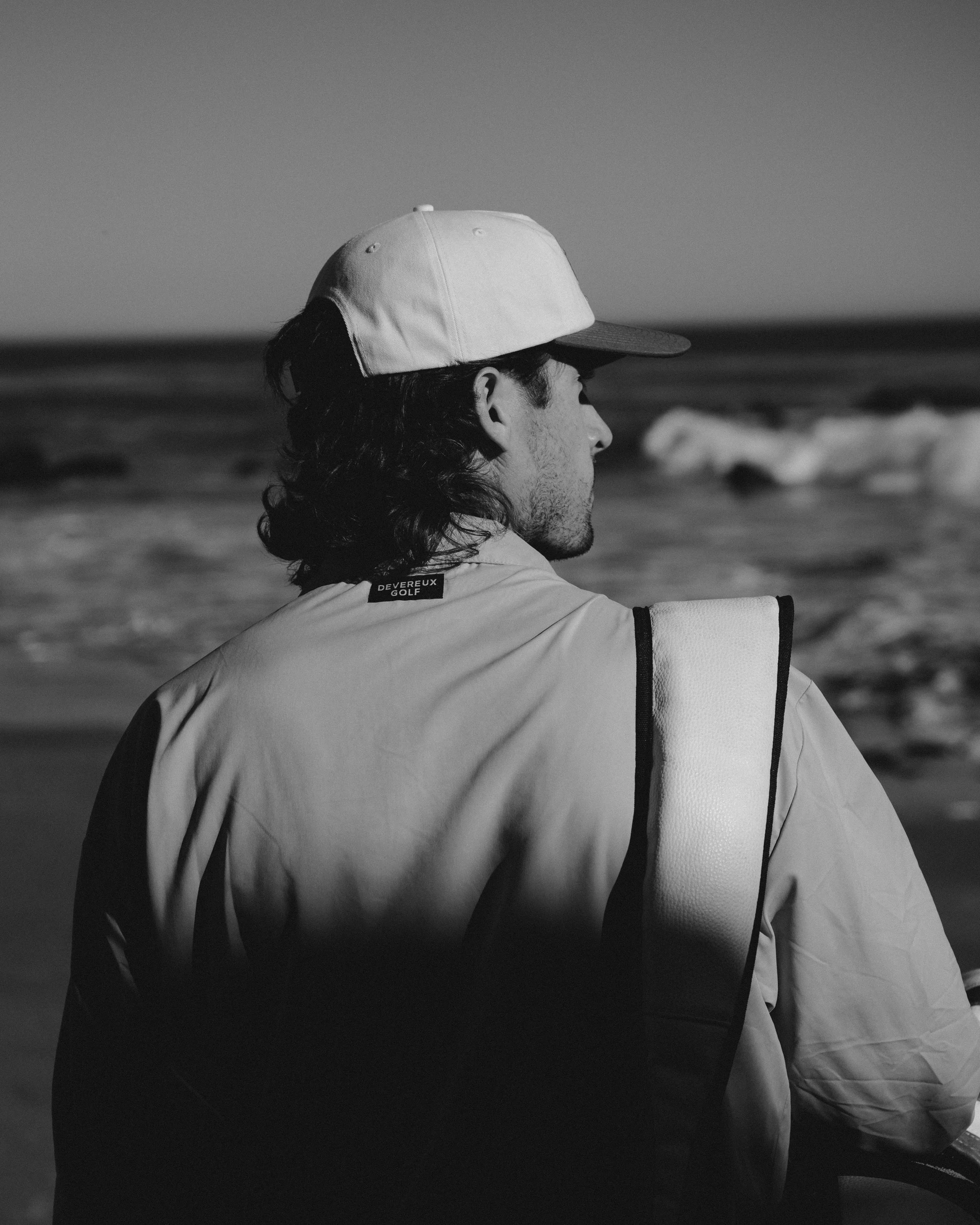 A man with long hair and a cap sitting on a beach, looking at the ocean waves.