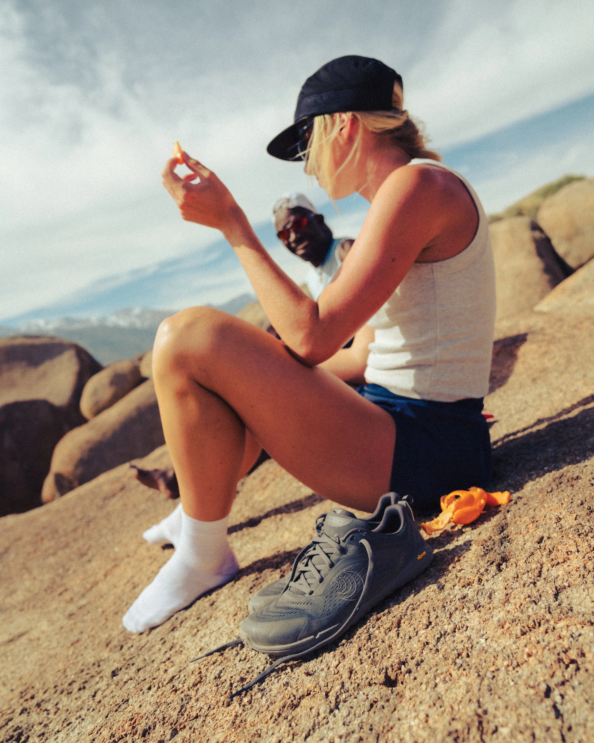 A woman sitting on a sandy beach, wearing a baseball cap and tank top, holding a snack, with a man in sunglasses and a cap in the background. There are rocks and mountains in the distance.