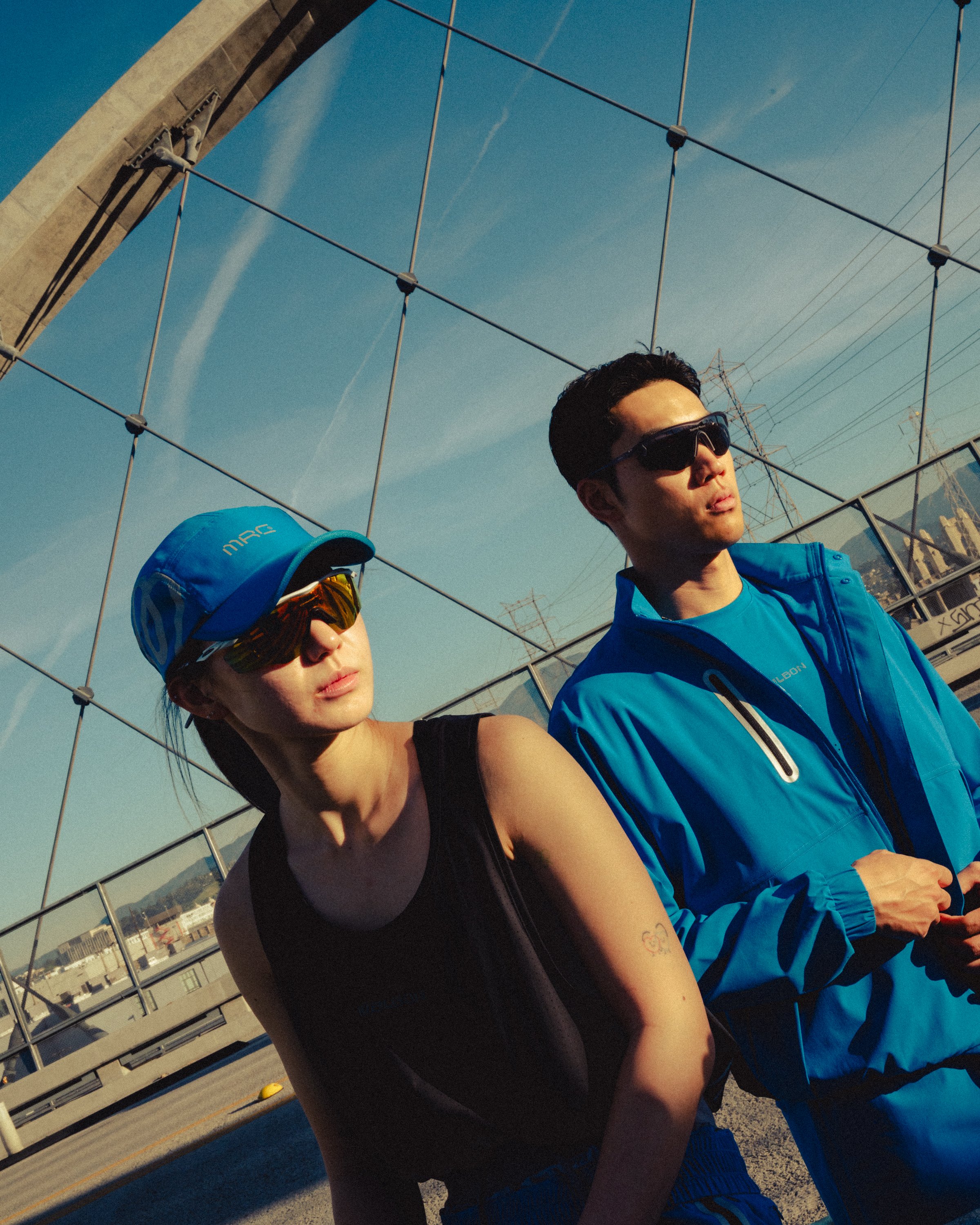 Two people wearing sports gear and sunglasses stand outdoors next to a chain-link fence, with an industrial background and blue sky.