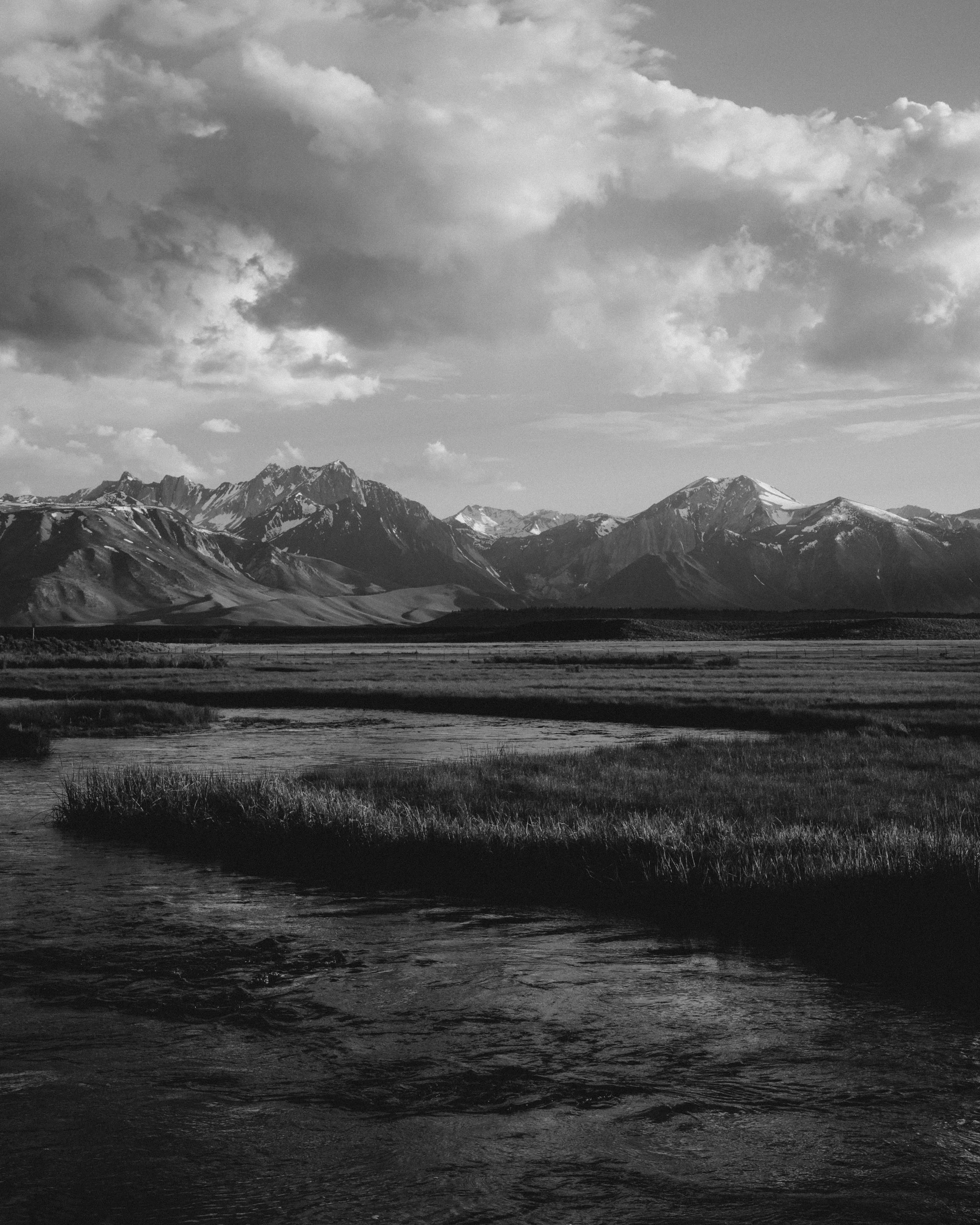 A black and white landscape featuring a river in the foreground, a range of snow-capped mountains in the background, and a sky filled with clouds.