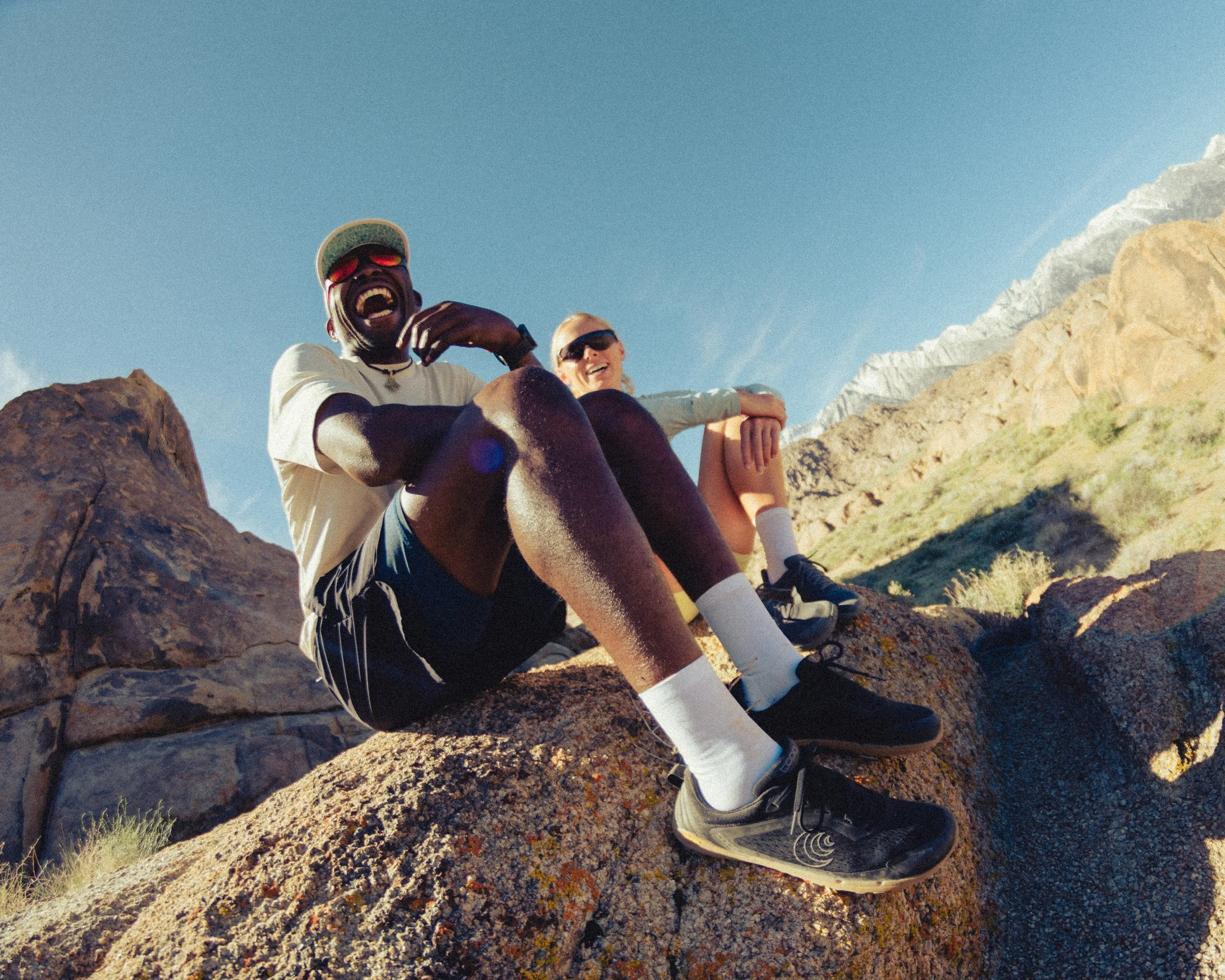 Two people sitting on rocks outdoors with mountainous terrain in the background, smiling at the camera.
