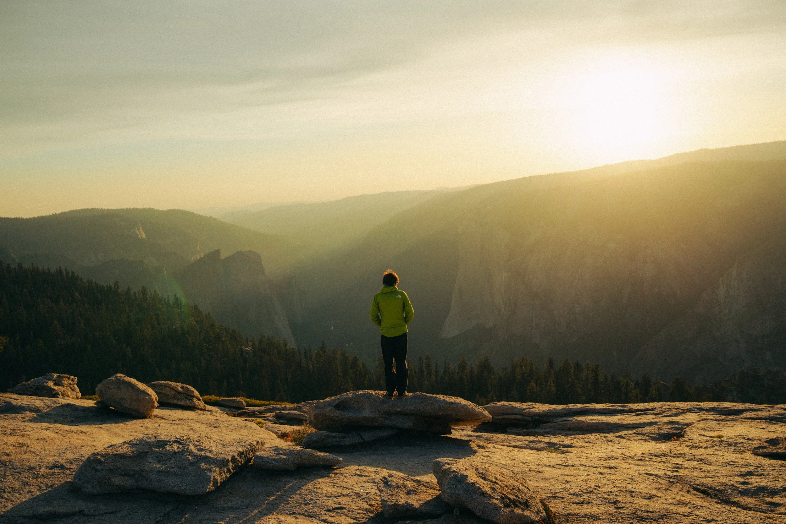 A person in a green jacket standing on rocky terrain, overlooking a valley with mountains in the distance during sunset or sunrise.