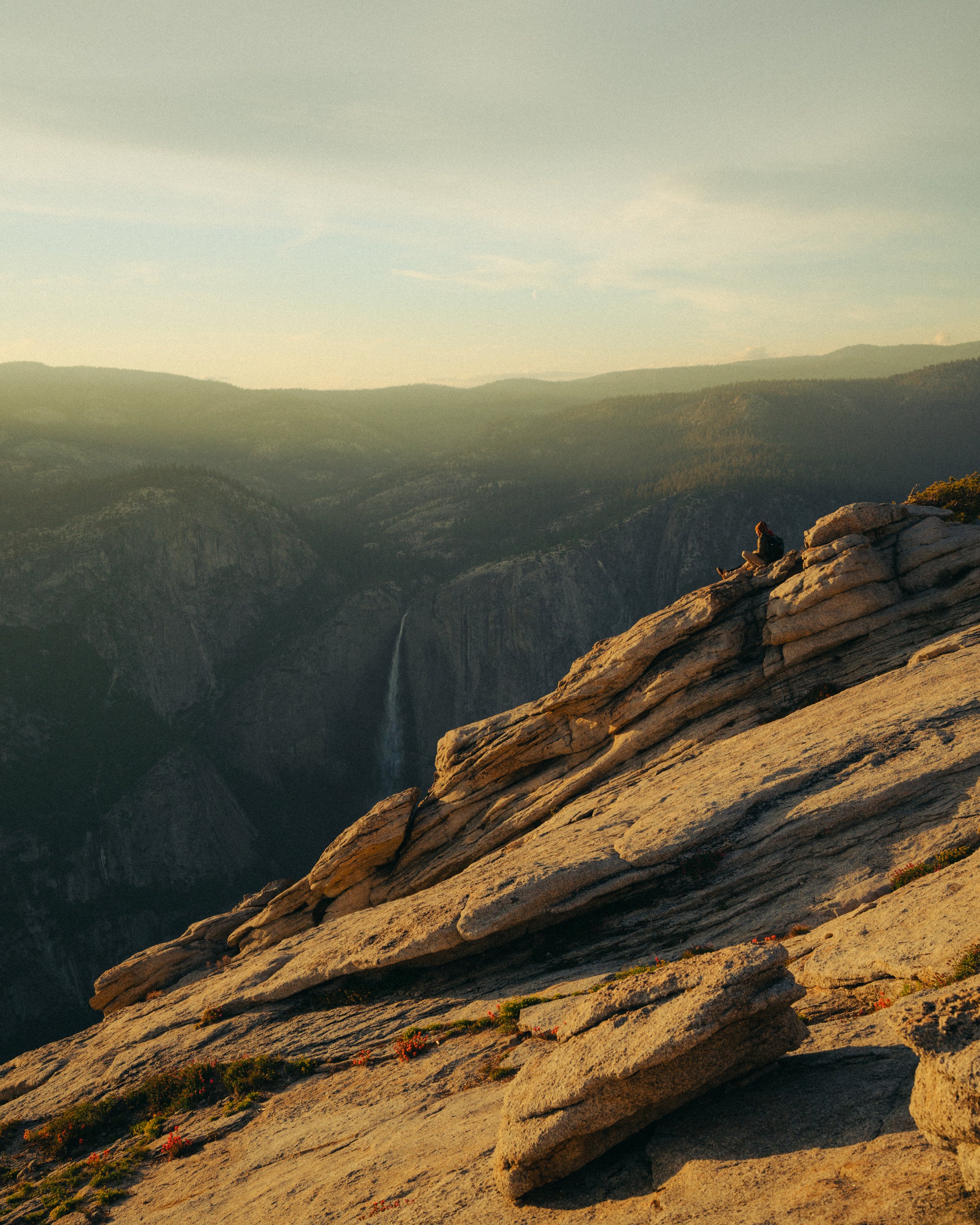 A person sitting on a large rock on a mountain edge, overlooking a deep valley with lush green ridges, during sunset or sunrise.