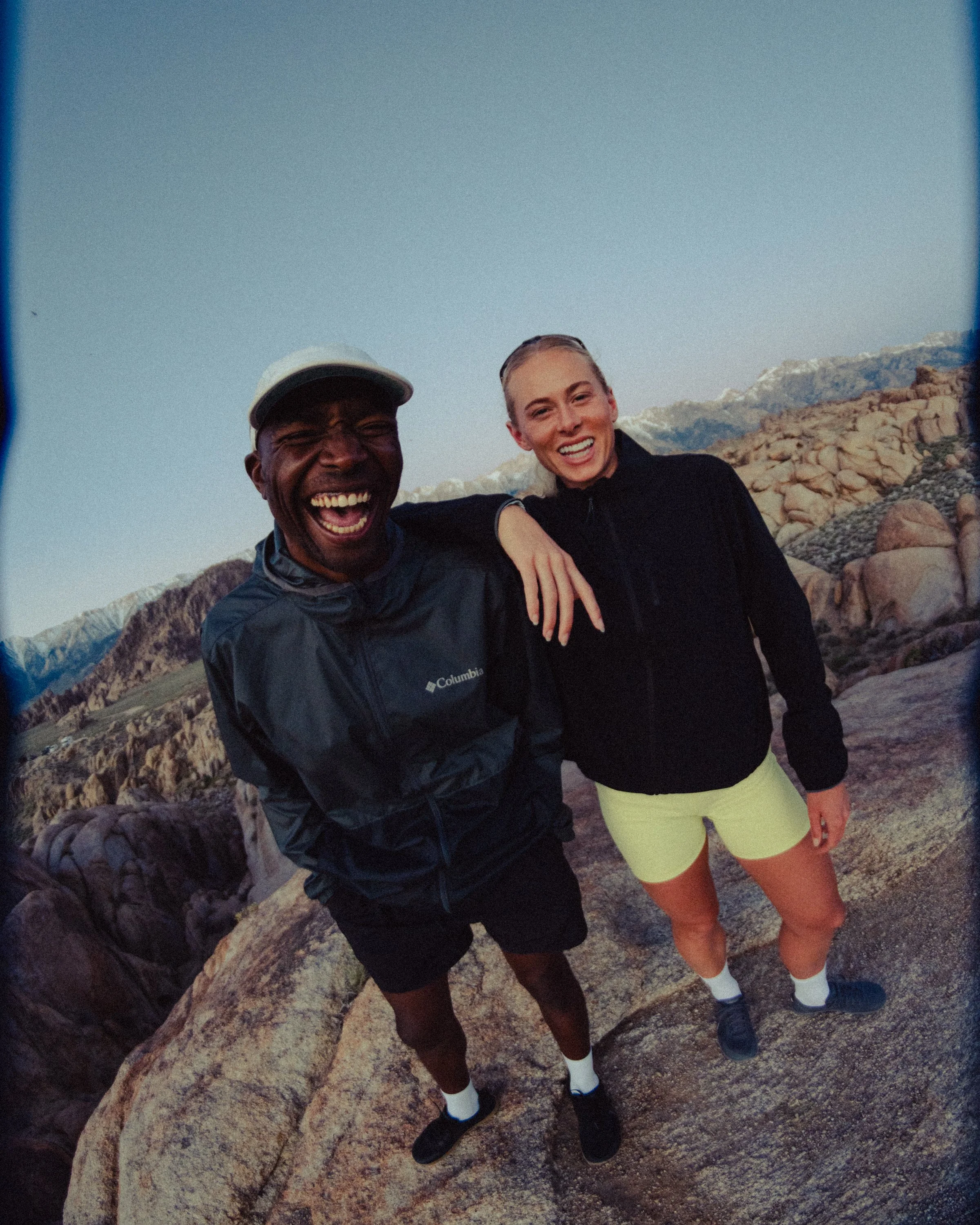 Two friends smiling at the camera, standing on large rocks in a mountainous outdoor setting during sunset or sunrise.