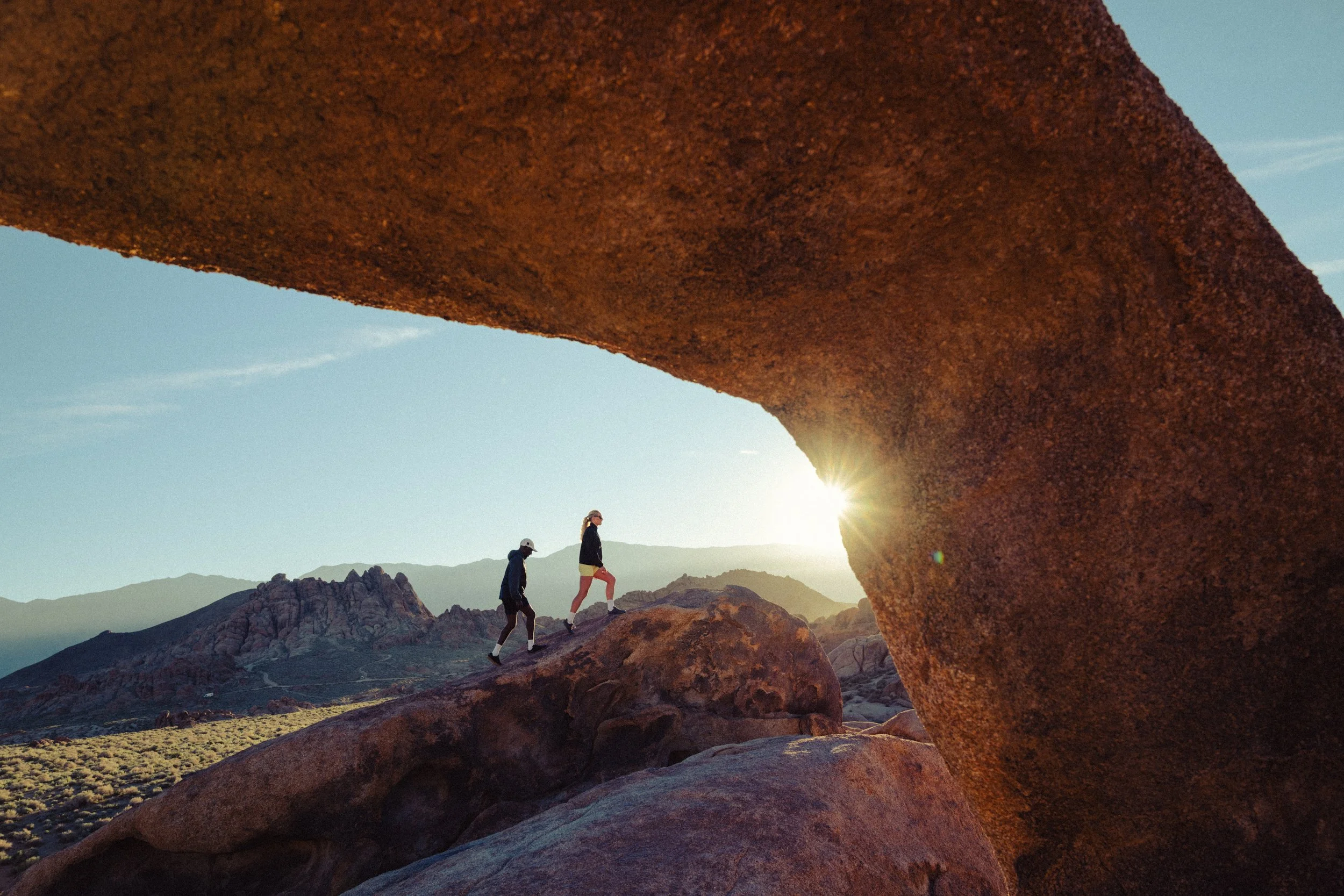 Three hikers walking through a rocky desert landscape at sunset, with a large rock formation arching overhead and distant mountains in the background.