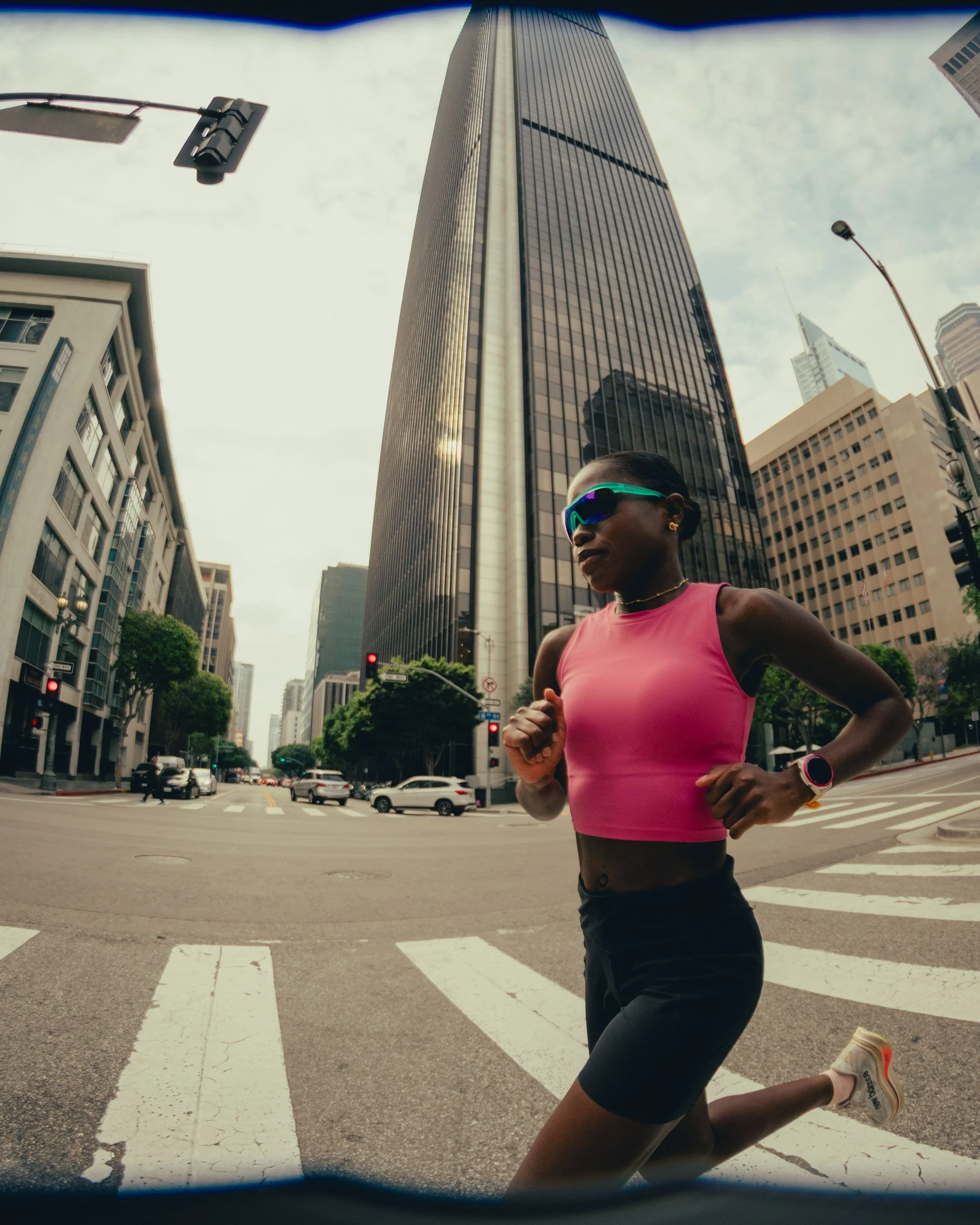 A woman running on a city street, wearing a pink athletic top, black shorts, sunglasses, and a smartwatch, with tall modern buildings in the background.