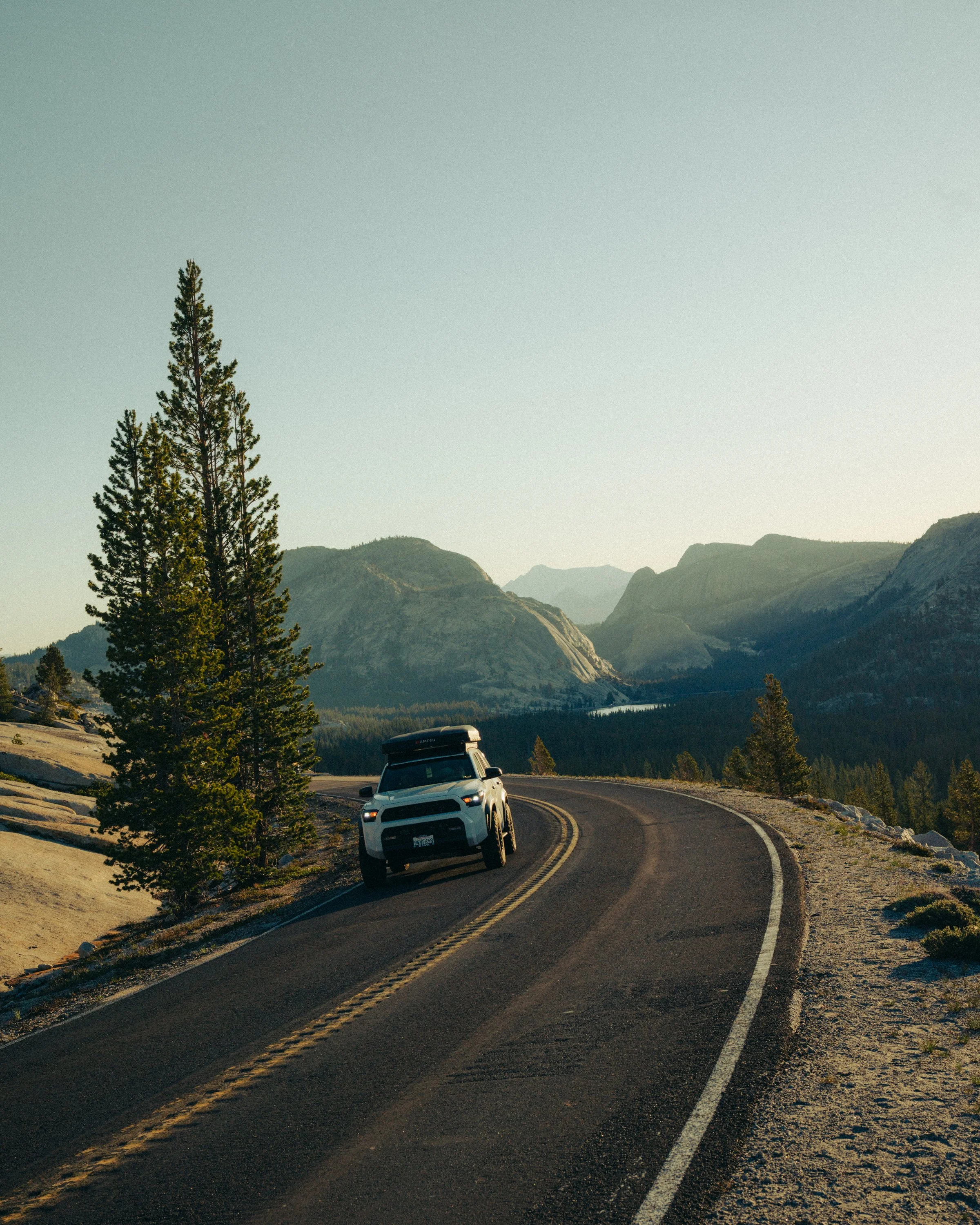 A winding mountain road with a white vehicle driving through a scenic landscape of trees and mountains at sunset or sunrise.