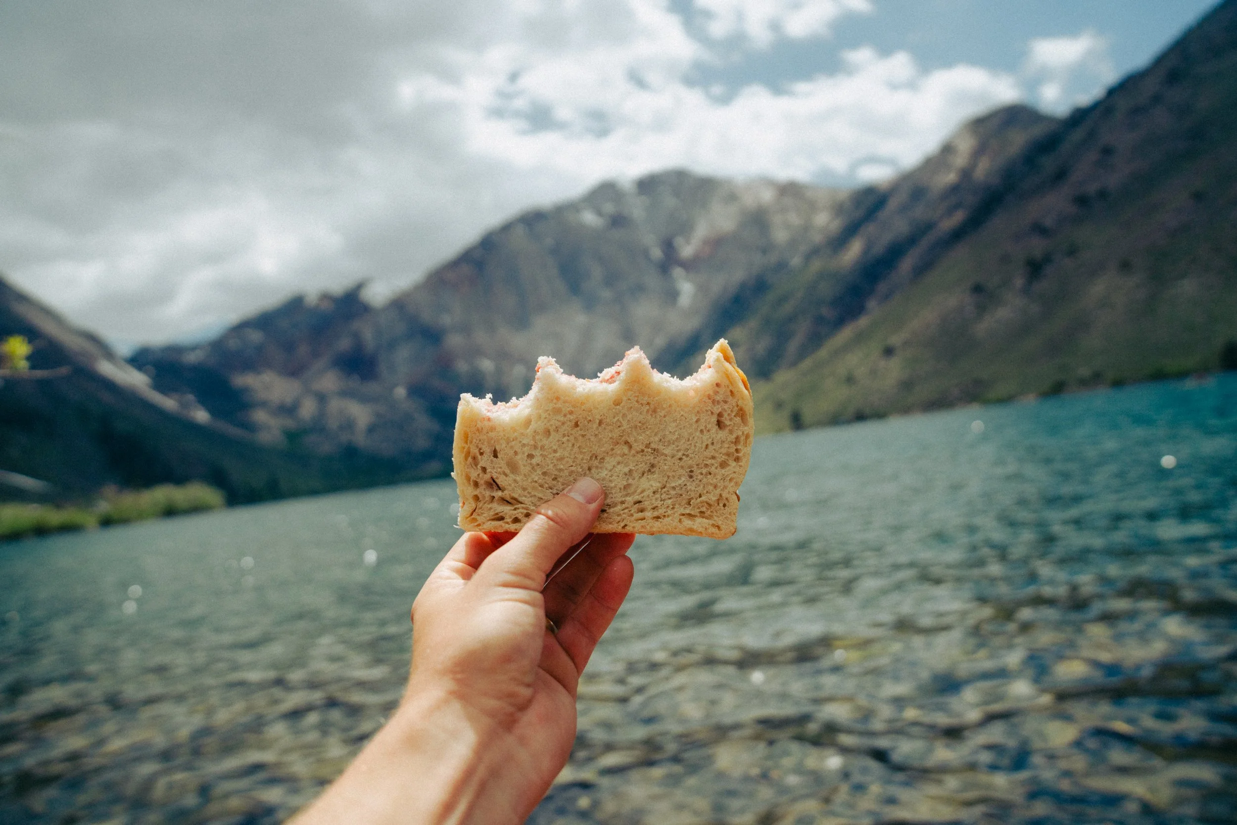 A person holding a slice of bread in front of a mountain landscape with water and cloudy sky.